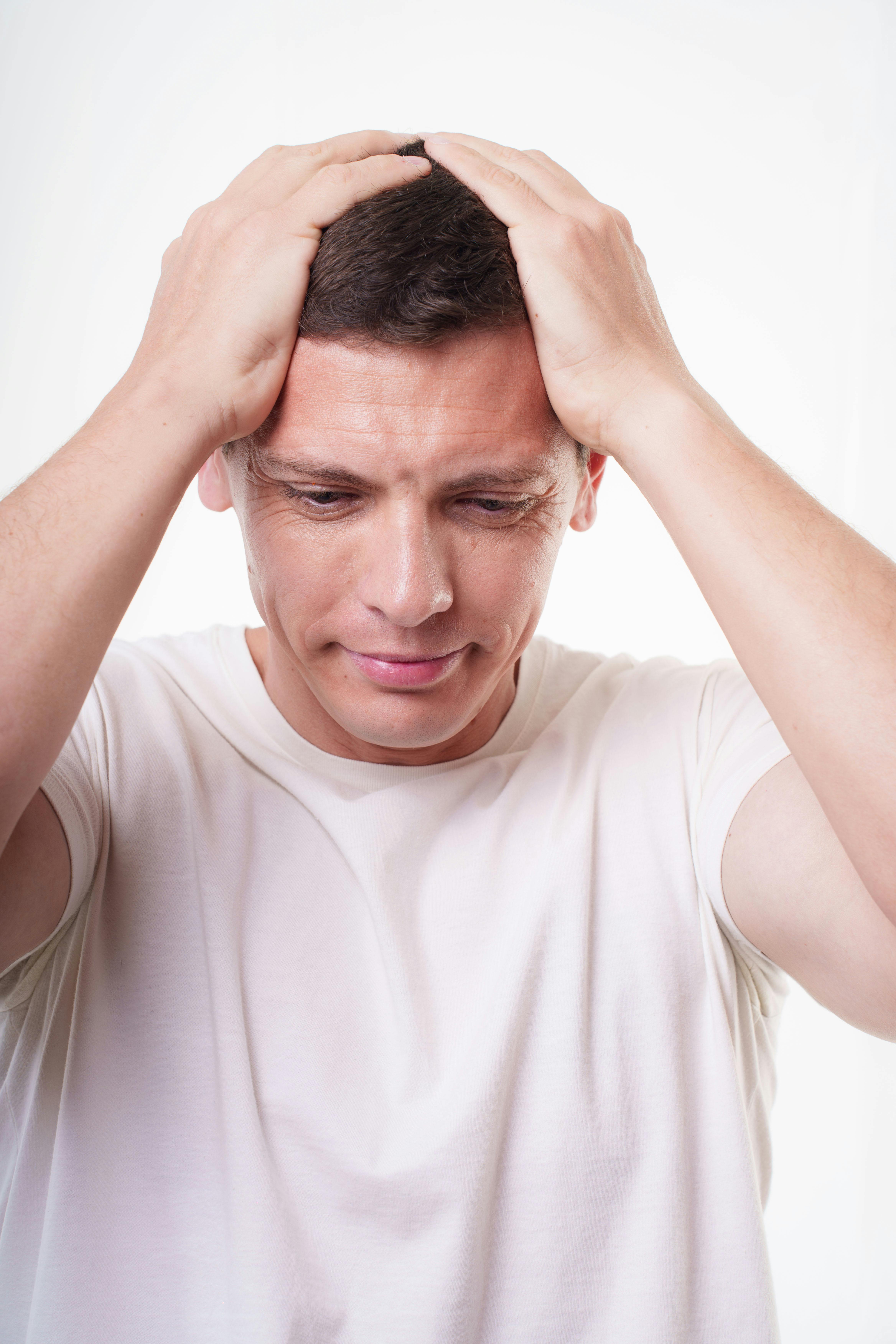 Free A young adult man in a white shirt appears stressed, holding his head with both hands against a plain background. Stock Photo