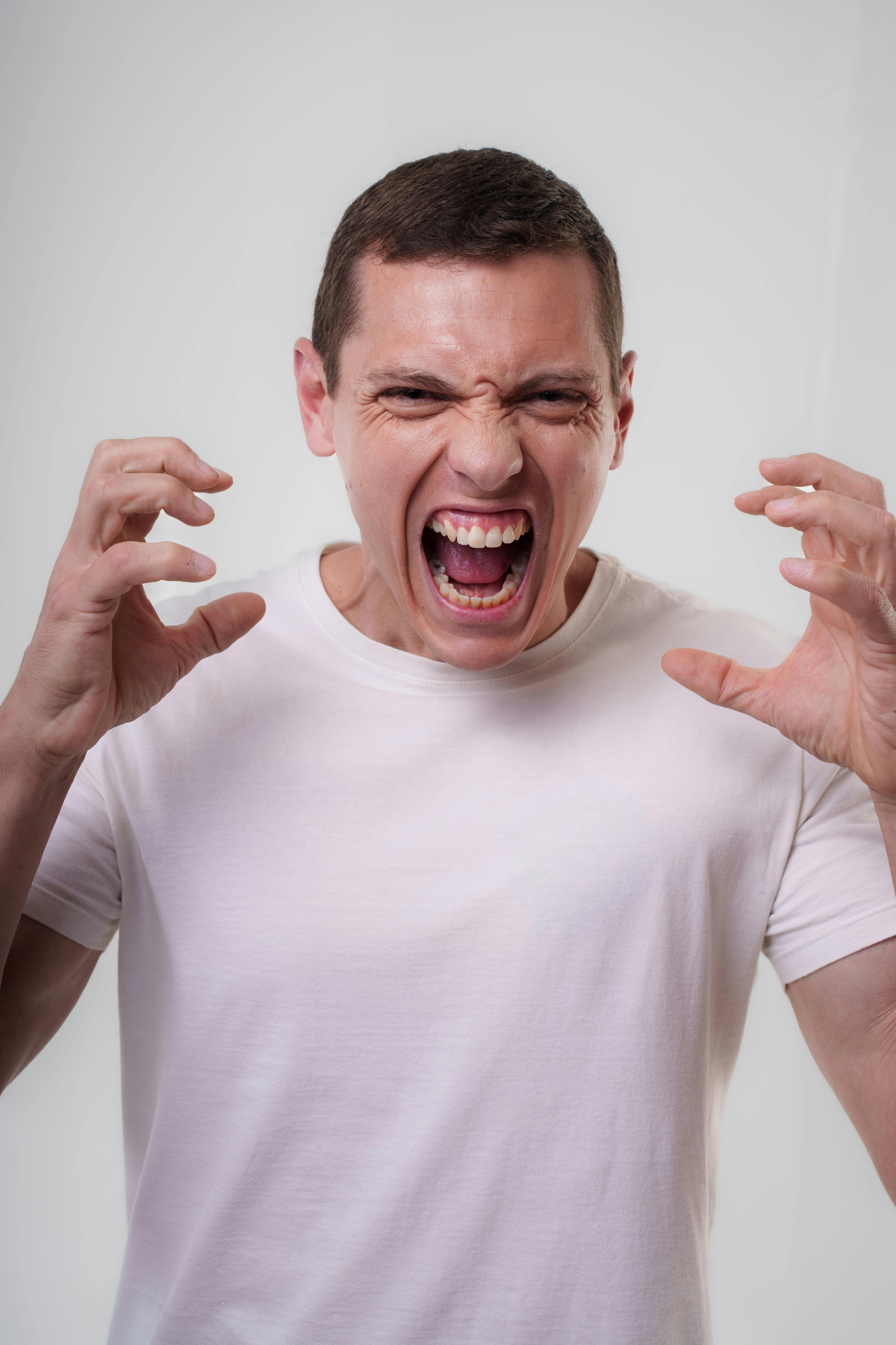 An adult man in a white t-shirt expressing strong emotions by shouting indoors.