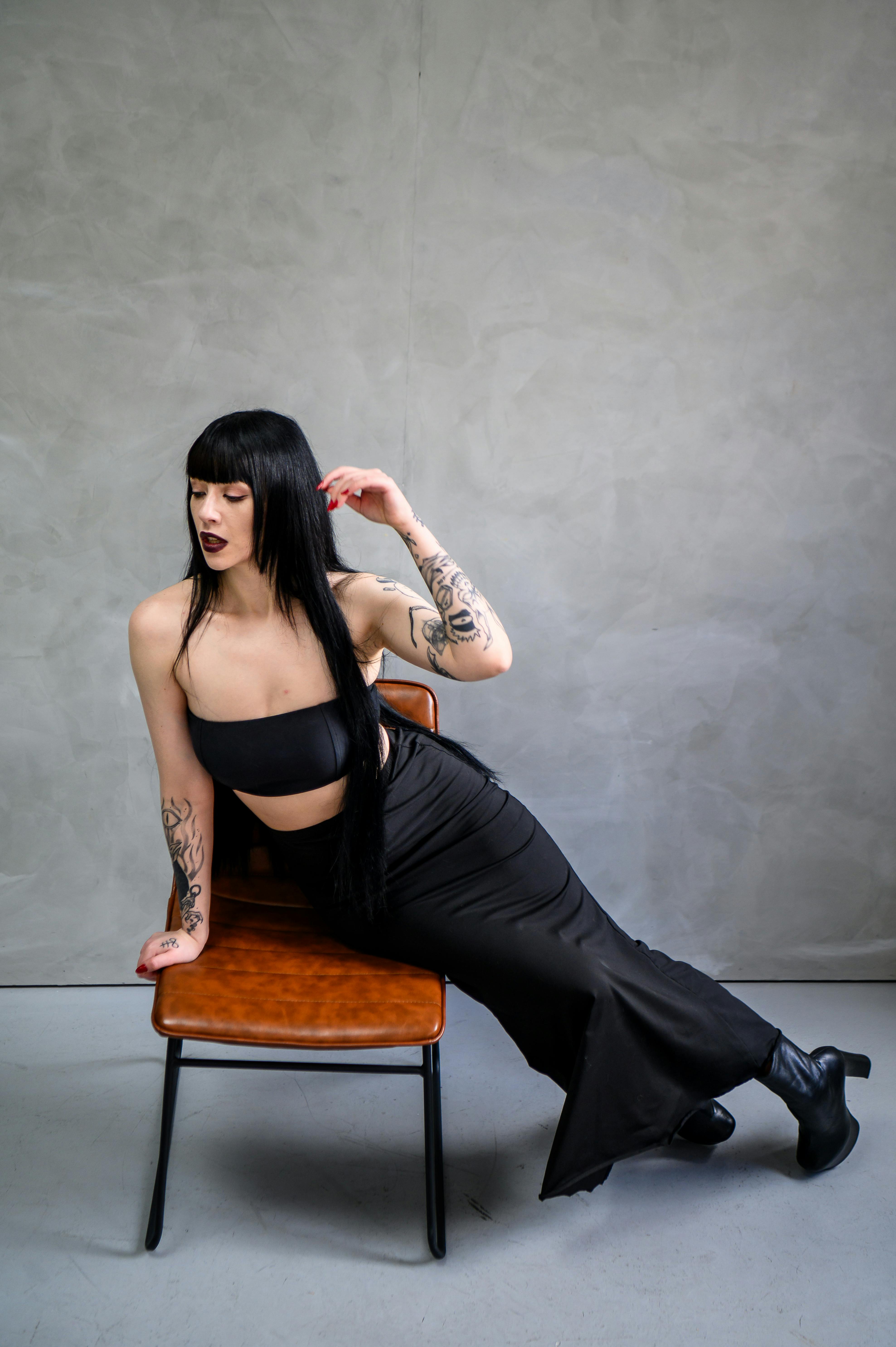 Bezpłatne Stylowa kobieta w czarnej sukience pozuje na krześle w nowoczesnym studiu. Zdjęcie z galerii