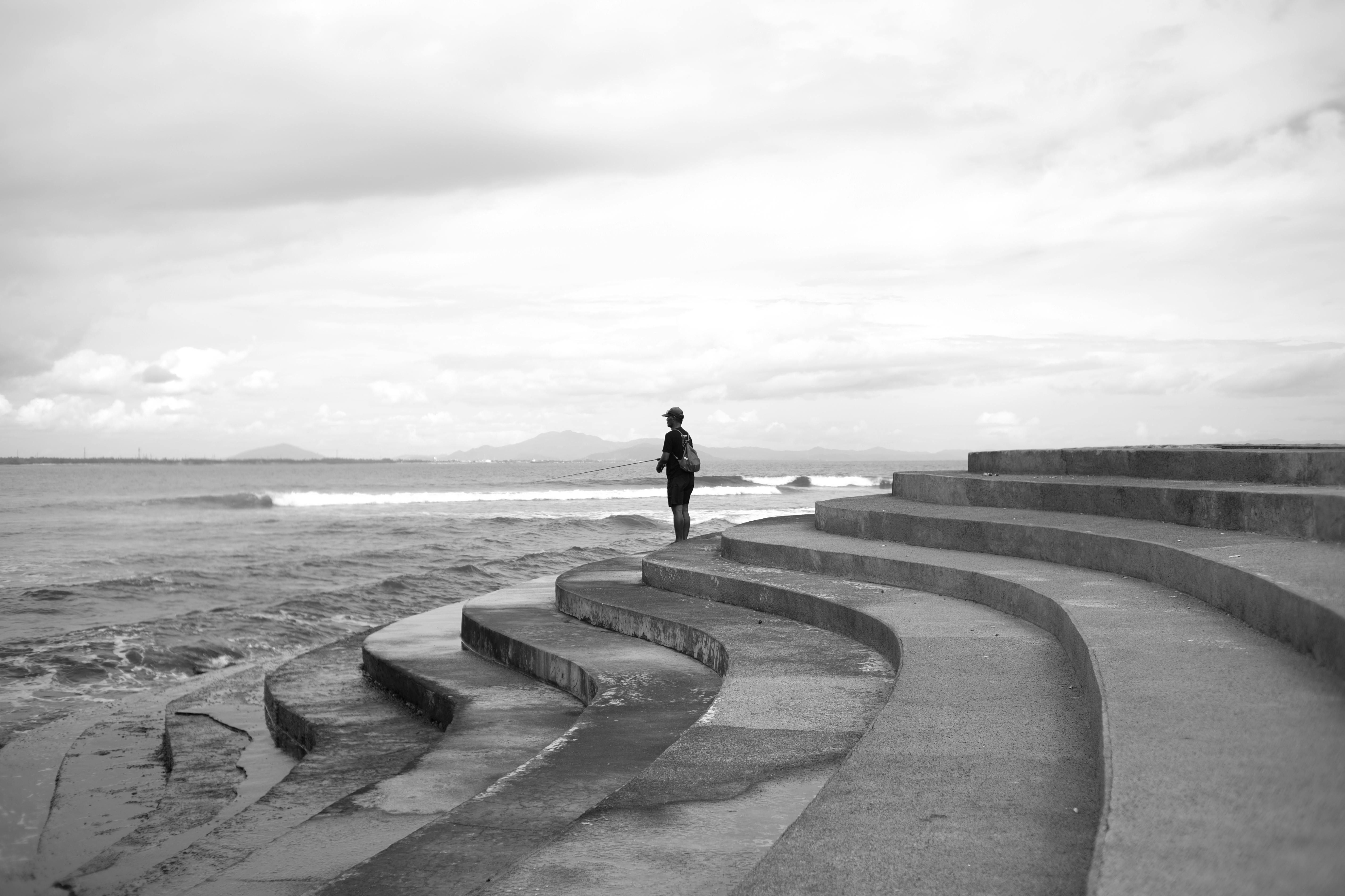 Black and white image of a person walking by the ocean on curved concrete steps.