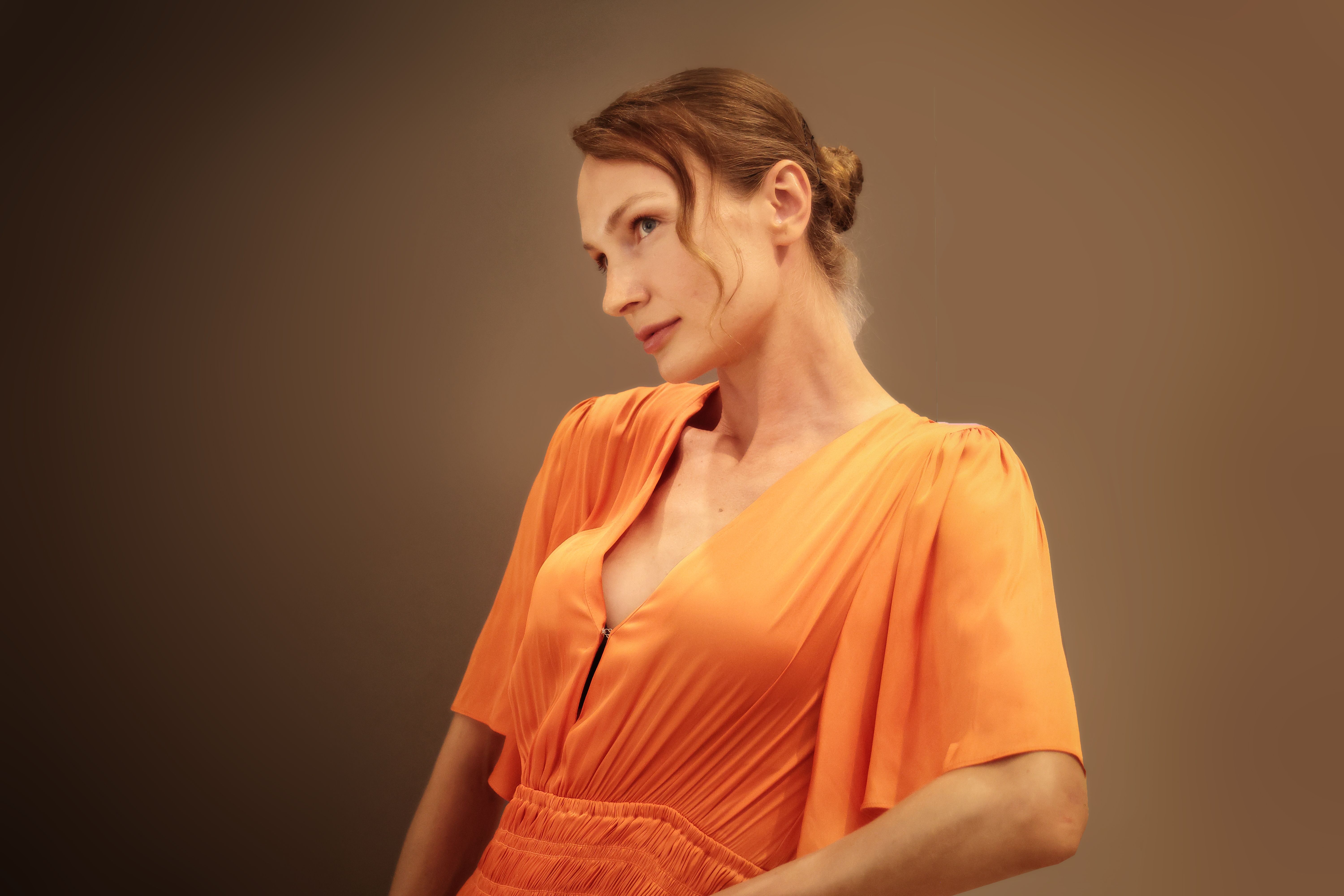 Une femme vêtue d'une robe orange pose avec style sur un fond marron doux avec un éclairage élégant.
