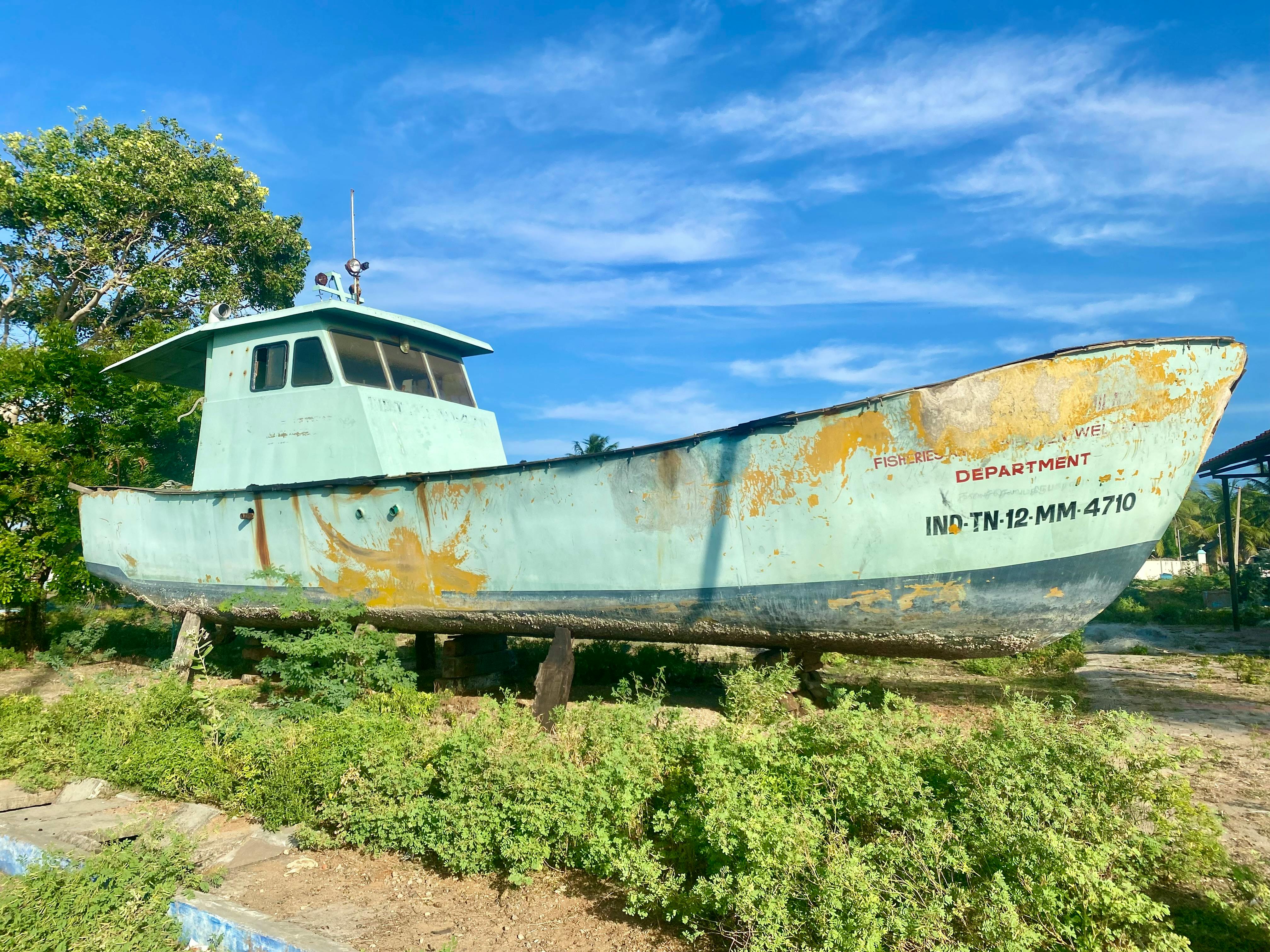 A weathered, old fishing boat resting on land surrounded by greenery, under a bright blue sky.