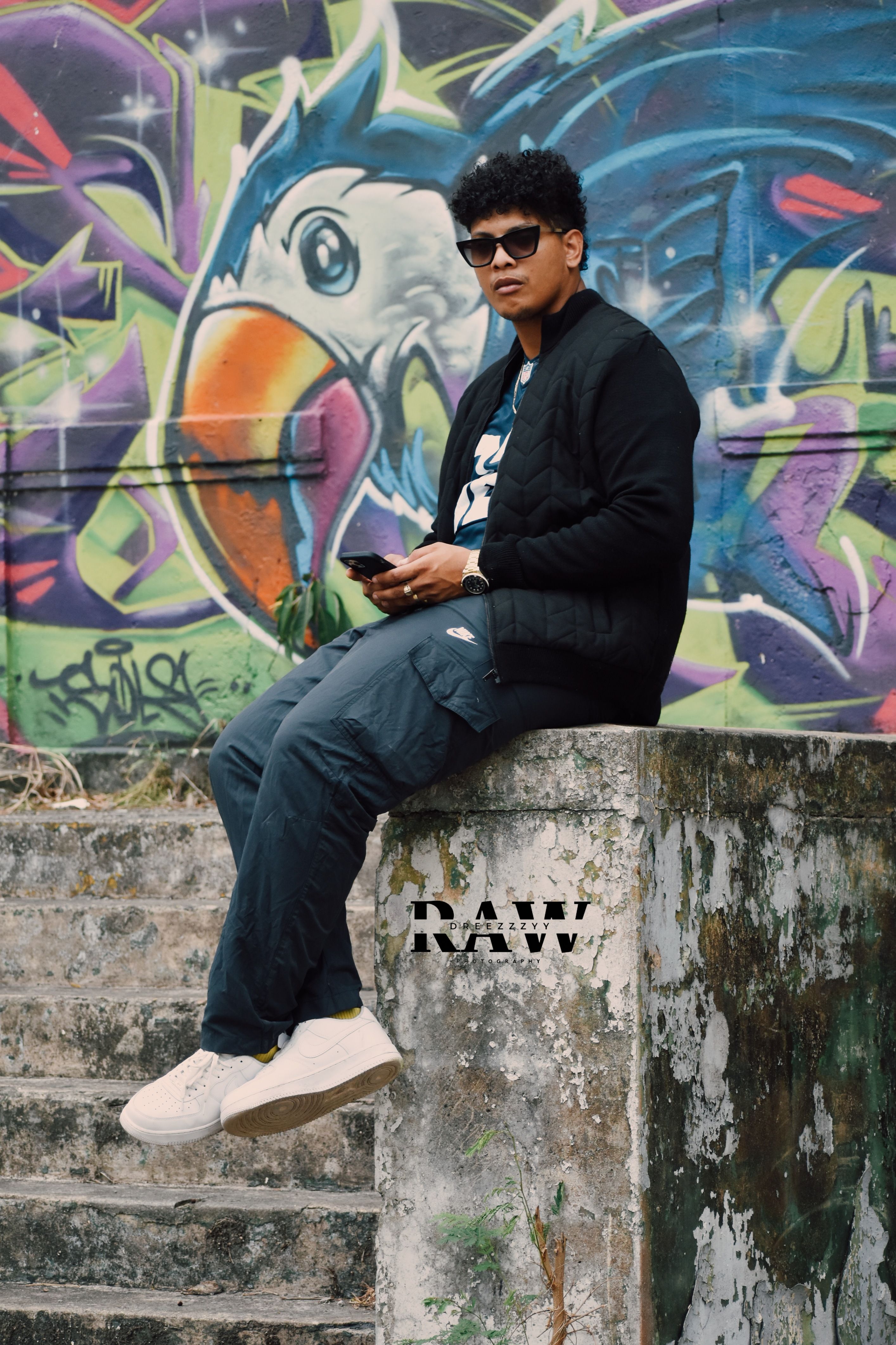 Young man sitting on steps against colorful graffiti mural outdoors.