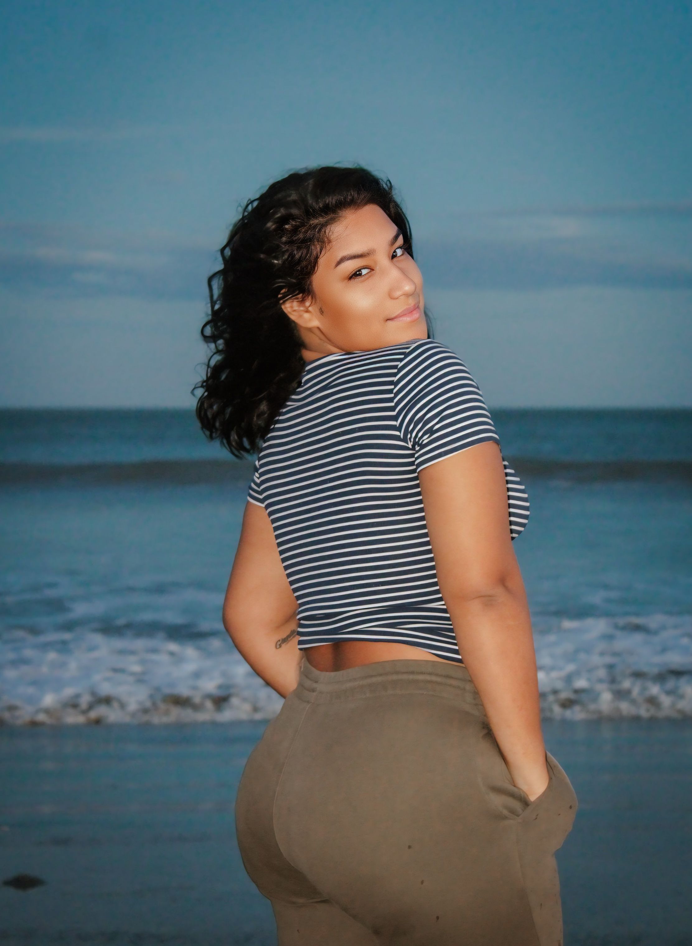 Young woman standing on the beach at sunset, gazing over her shoulder with a smile.