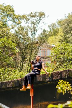 A young woman holding a camera sits on a rustic structure in a vibrant forest in Ocoyoacac, Mexico.