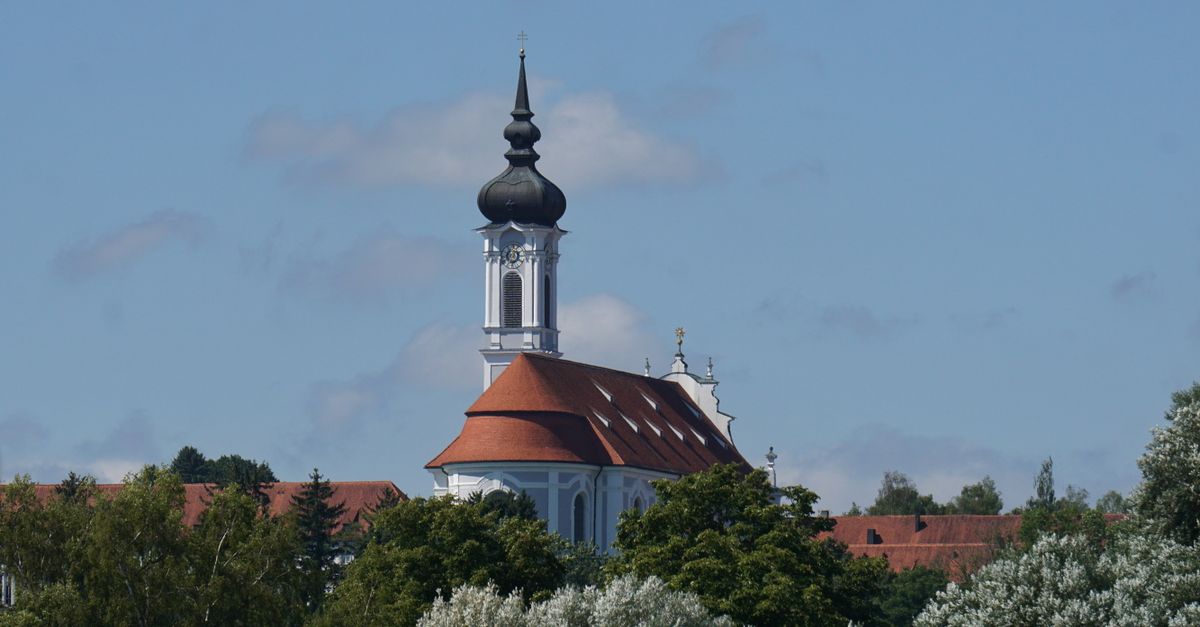 A picturesque view of a Baroque-style church tower surrounded by lush greenery under a clear blue sky.