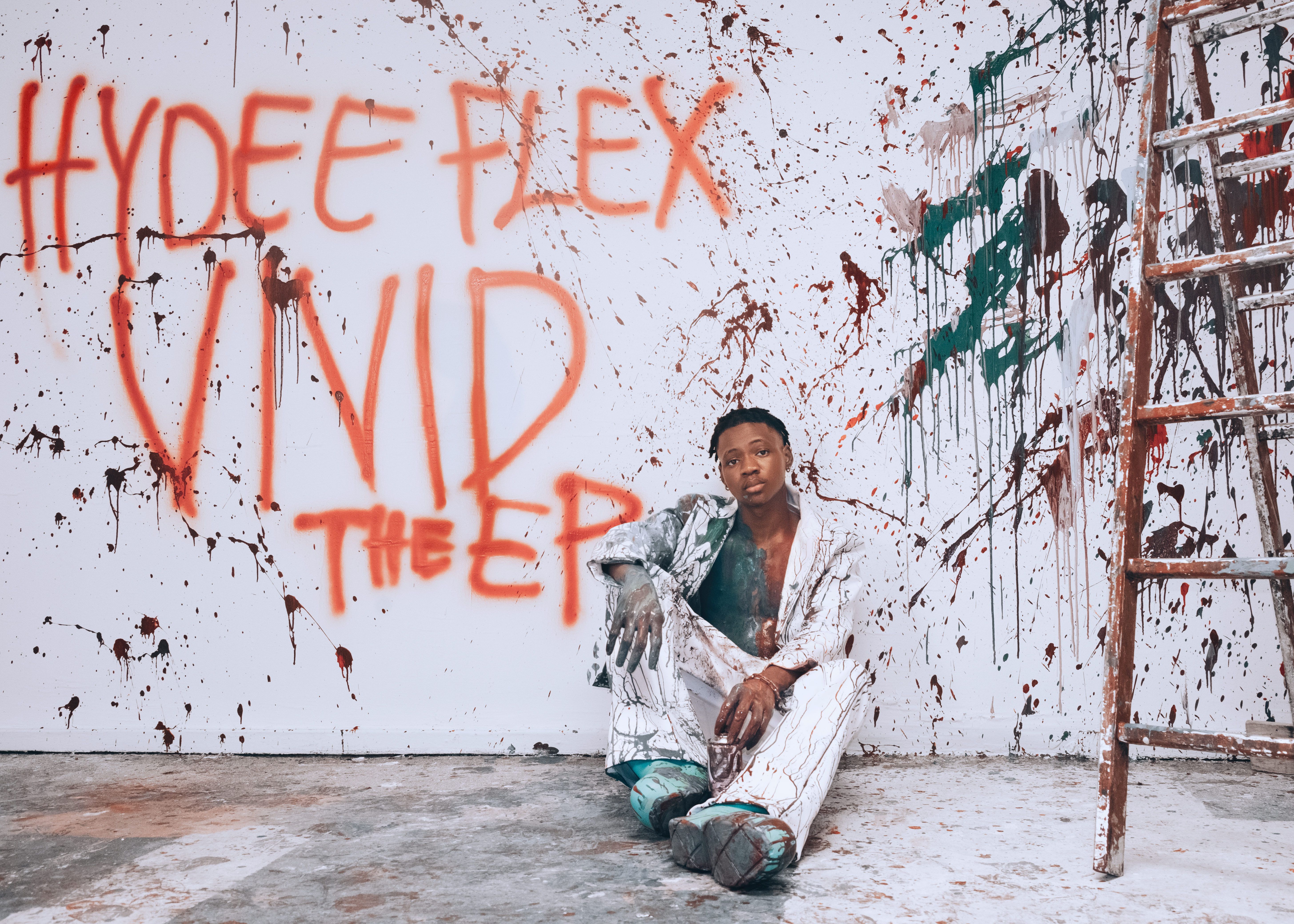 Artist sits in a paint-splattered studio, next to spray-painted text on the wall.