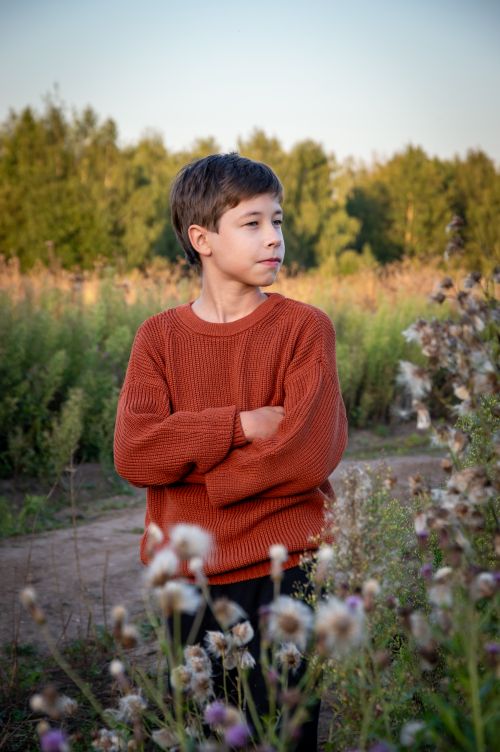 Free Boy in orange sweater standing in a field with wildflowers, looking thoughtful. Stock Photo