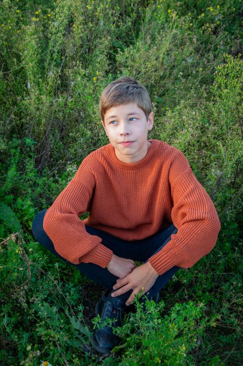 Free Young boy sitting in lush greenery, wearing a cozy orange sweater and black pants. Stock Photo