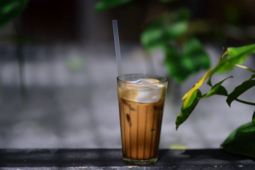Free Chilled iced coffee served in a tall glass with ice cubes and a straw, set outdoors with blurred greenery. Stock Photo