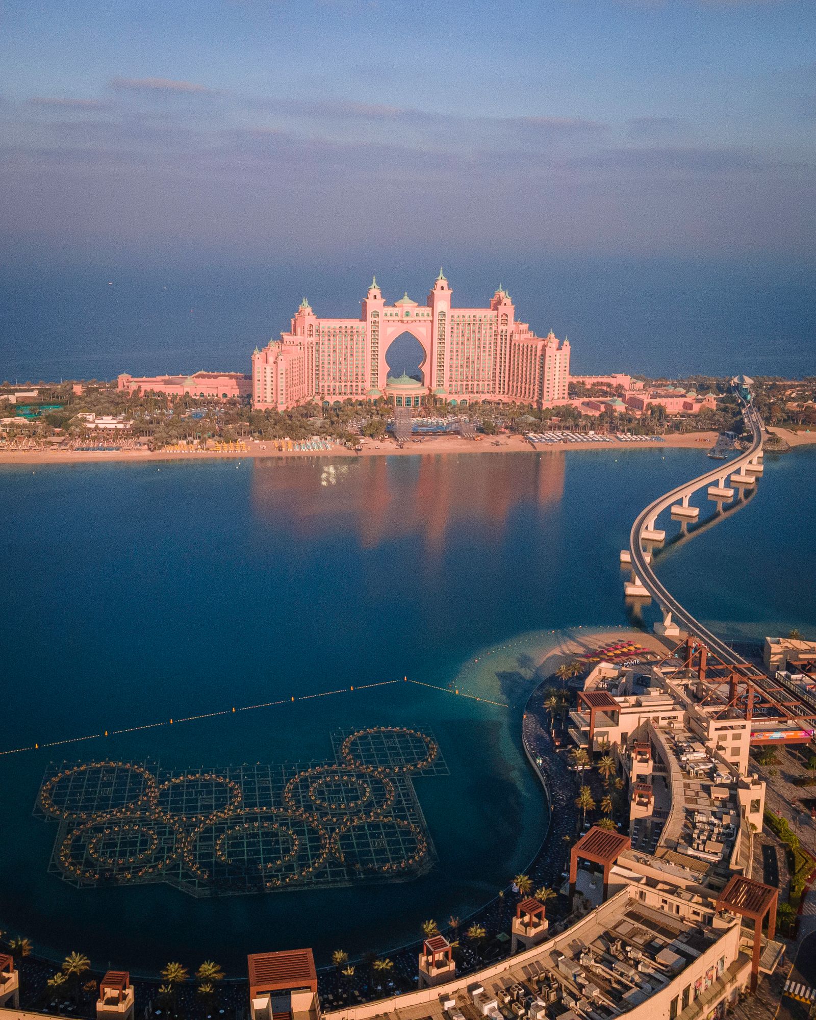 Atlantis The Palm at sunrise reflected in calm water, with the monorail causeway curving toward The Pointe on Palm Jumeirah