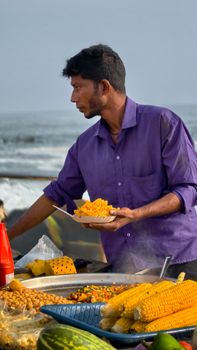 Man in a purple shirt serving street food at a beachside market with corn and pineapple.