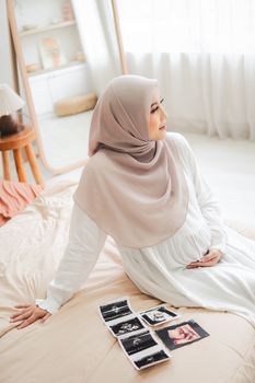 Expectant mother in hijab sitting in bedroom with ultrasound pictures, embracing motherhood. - Fotografer Sidoarjo