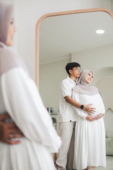 A loving couple embraces in front of a mirror, capturing a serene pregnancy moment. - Fotografer Sidoarjo