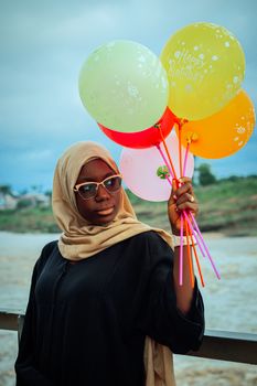 Stylish woman in hijab holds colorful birthday balloons outdoors.