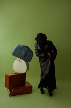 Fashionable individual in black coat and hat with vintage luggage on a green backdrop.