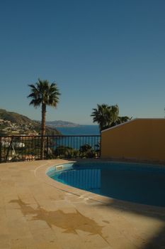 Serene view from a poolside overlooking the ocean with palm trees and distant mountains under a clear blue sky.