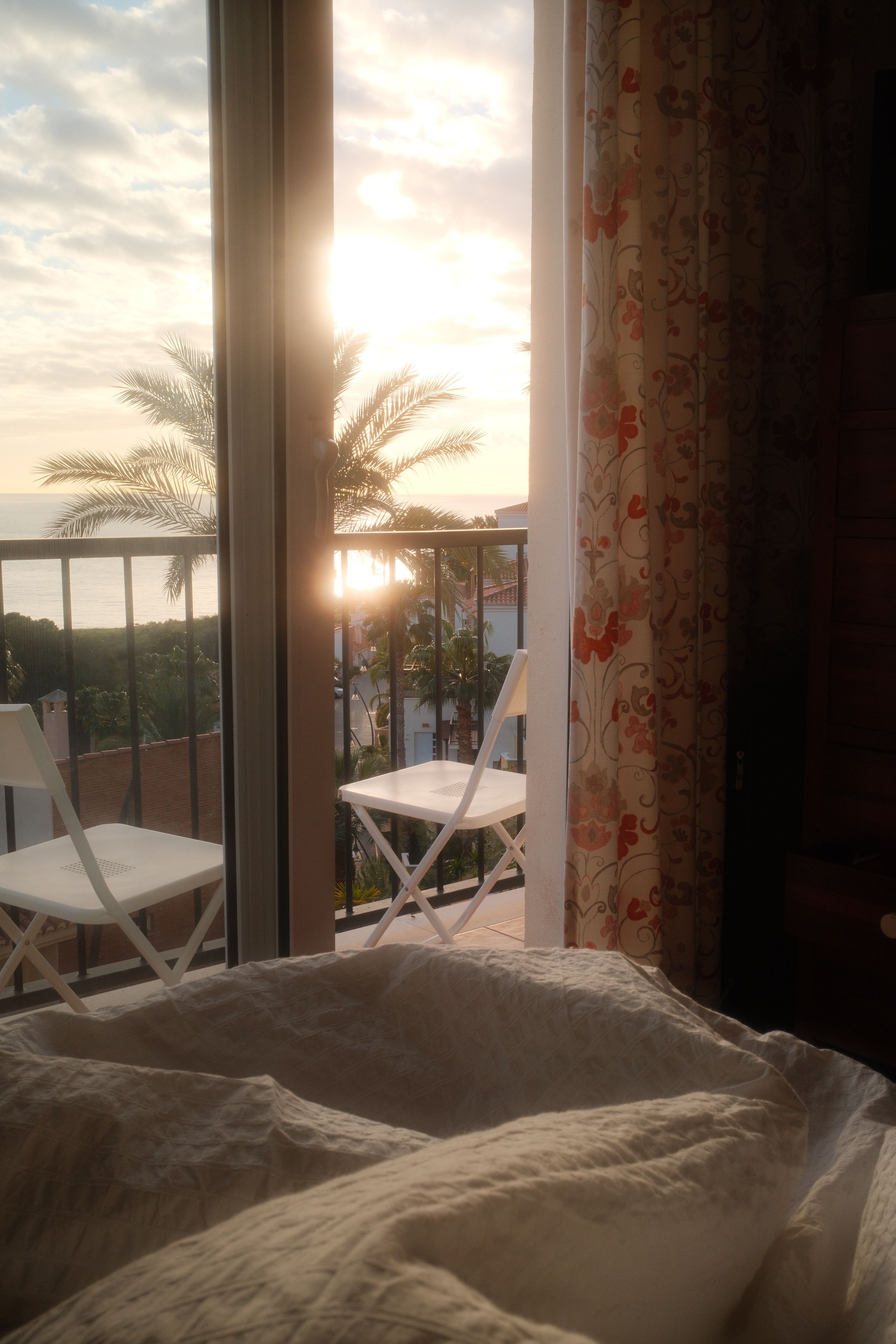 A tranquil sunrise view from a bedroom, overlooking a balcony with palm trees and ocean.