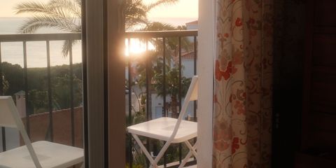 A tranquil sunrise view from a bedroom, overlooking a balcony with palm trees and ocean.