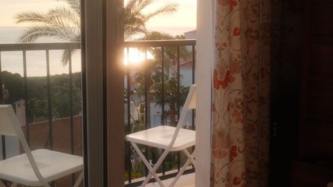 A tranquil sunrise view from a bedroom, overlooking a balcony with palm trees and ocean.