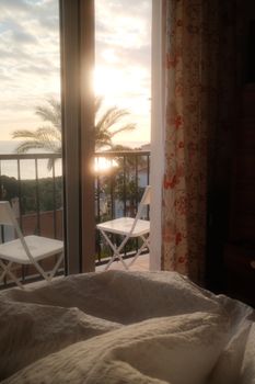 A tranquil sunrise view from a bedroom, overlooking a balcony with palm trees and ocean.