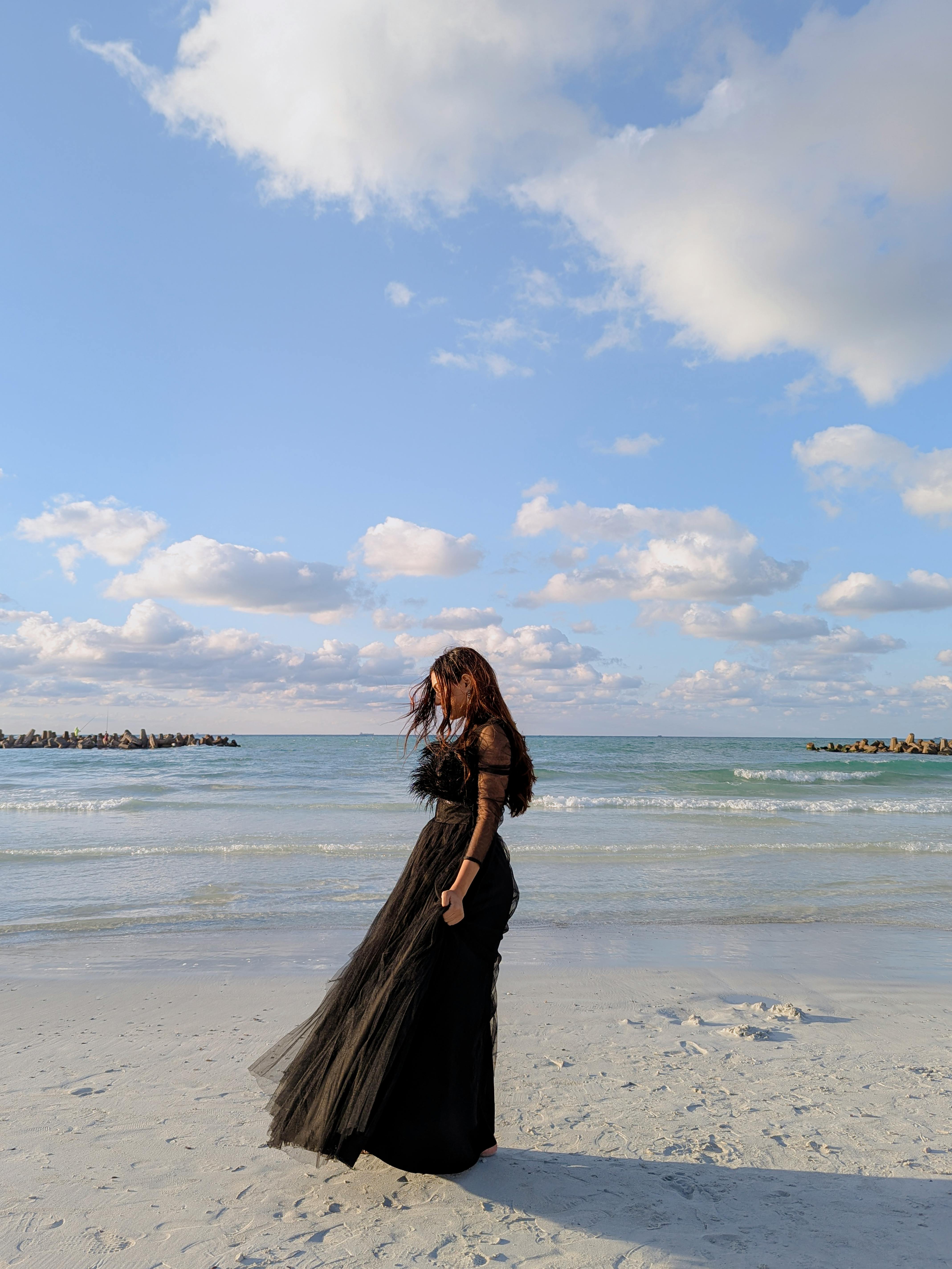 A woman in a black dress poses on a sunny beach with a scenic ocean backdrop.