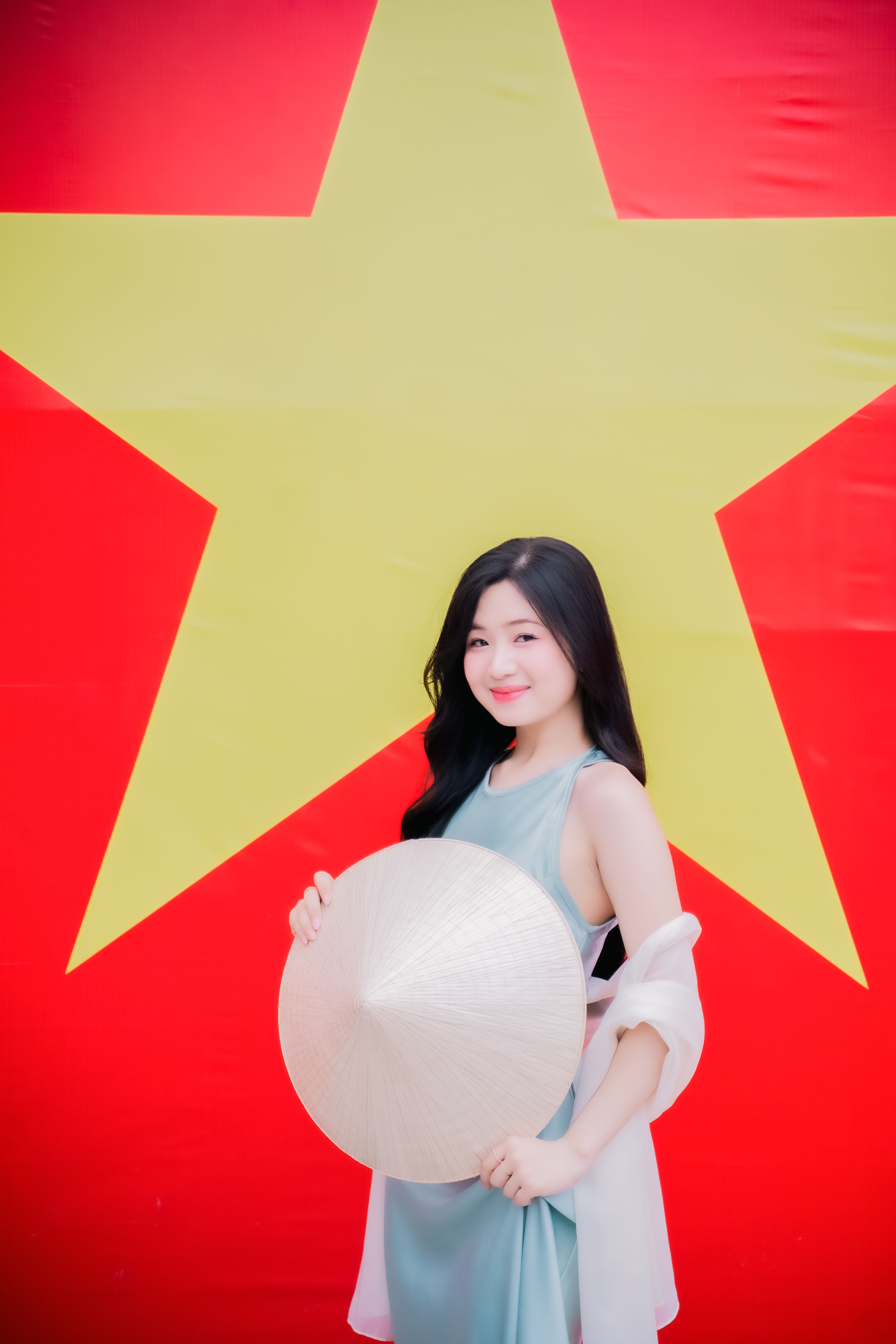 Young woman holding conical hat in front of Vietnamese flag, vibrant colors.