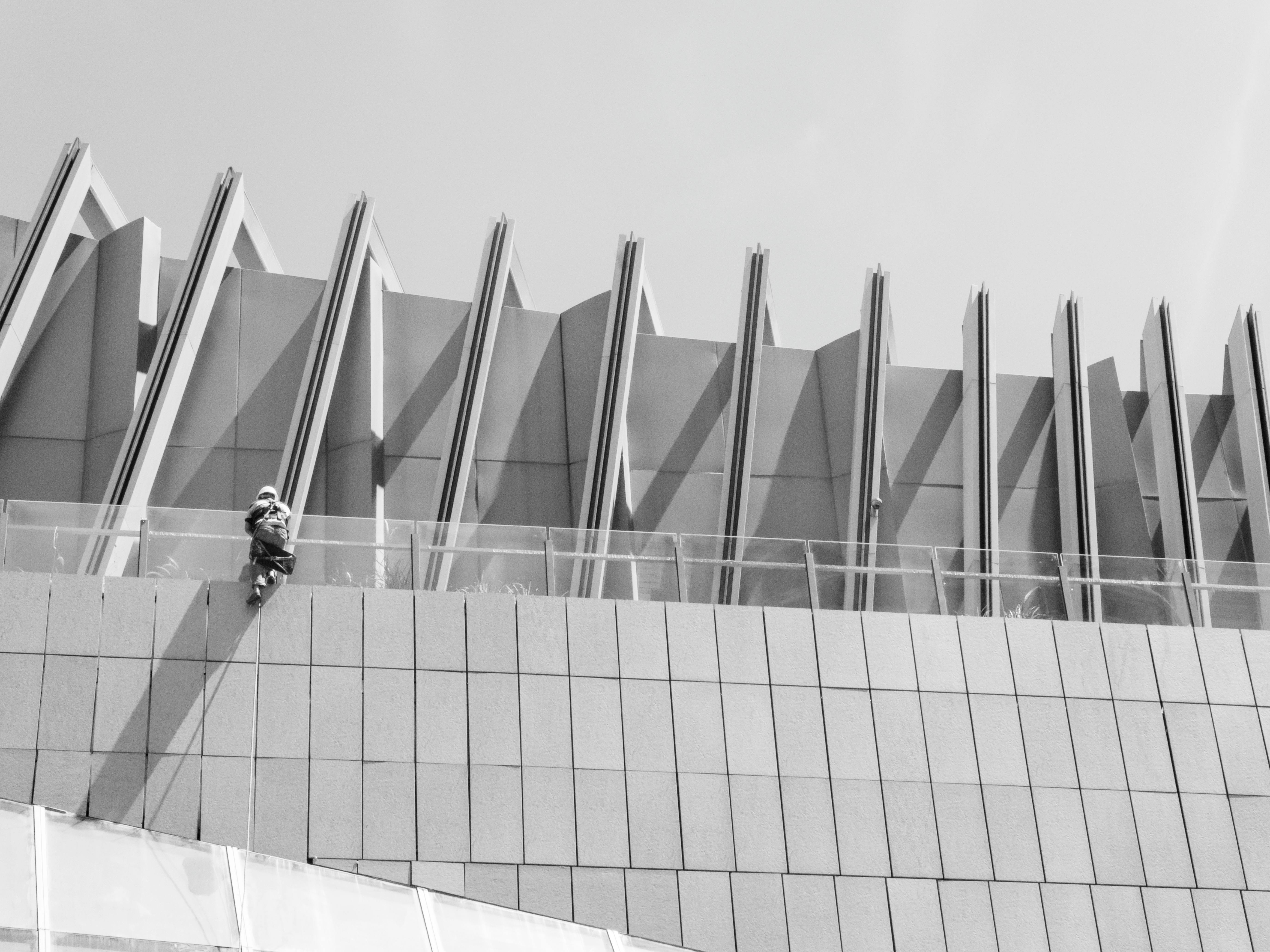 Gratis Foto in bianco e nero di un operaio impegnato nella manutenzione della facciata di un edificio moderno dalle linee architettoniche sorprendenti. Foto a disposizione