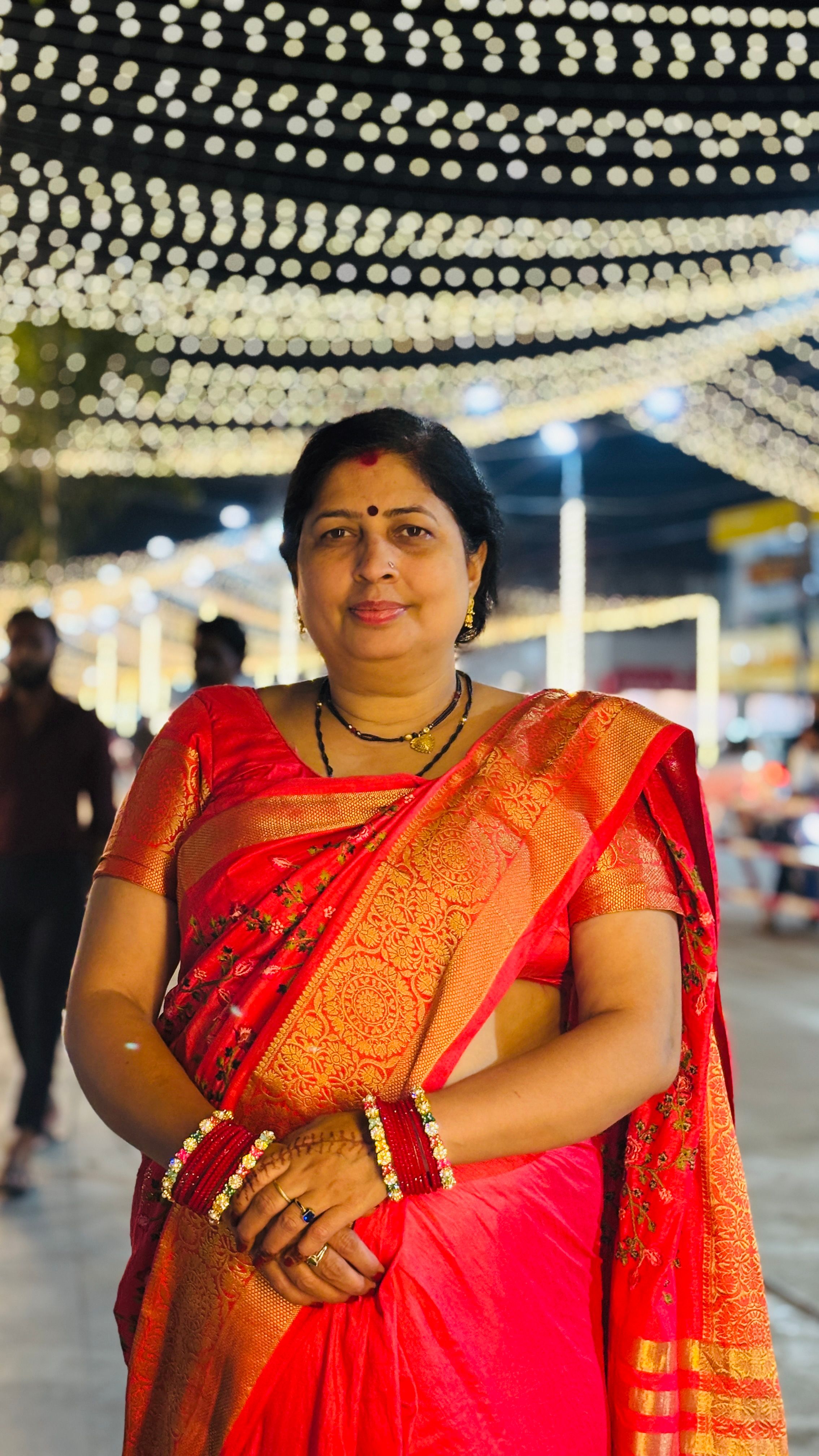 A smiling Indian woman in a vibrant red sari stands under festive lights at a night celebration.