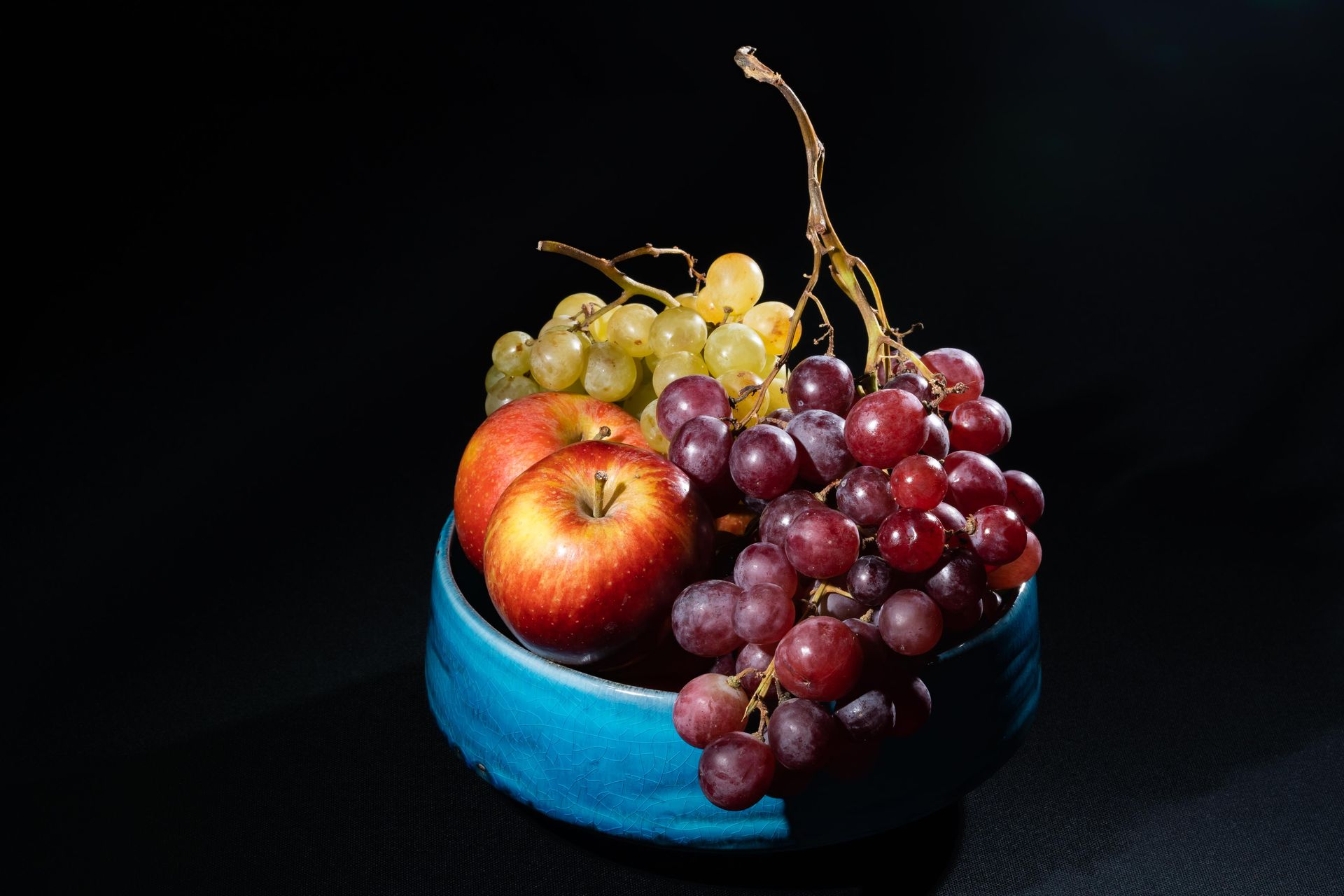Still life of apples and grapes in a vibrant blue bowl on a black background.