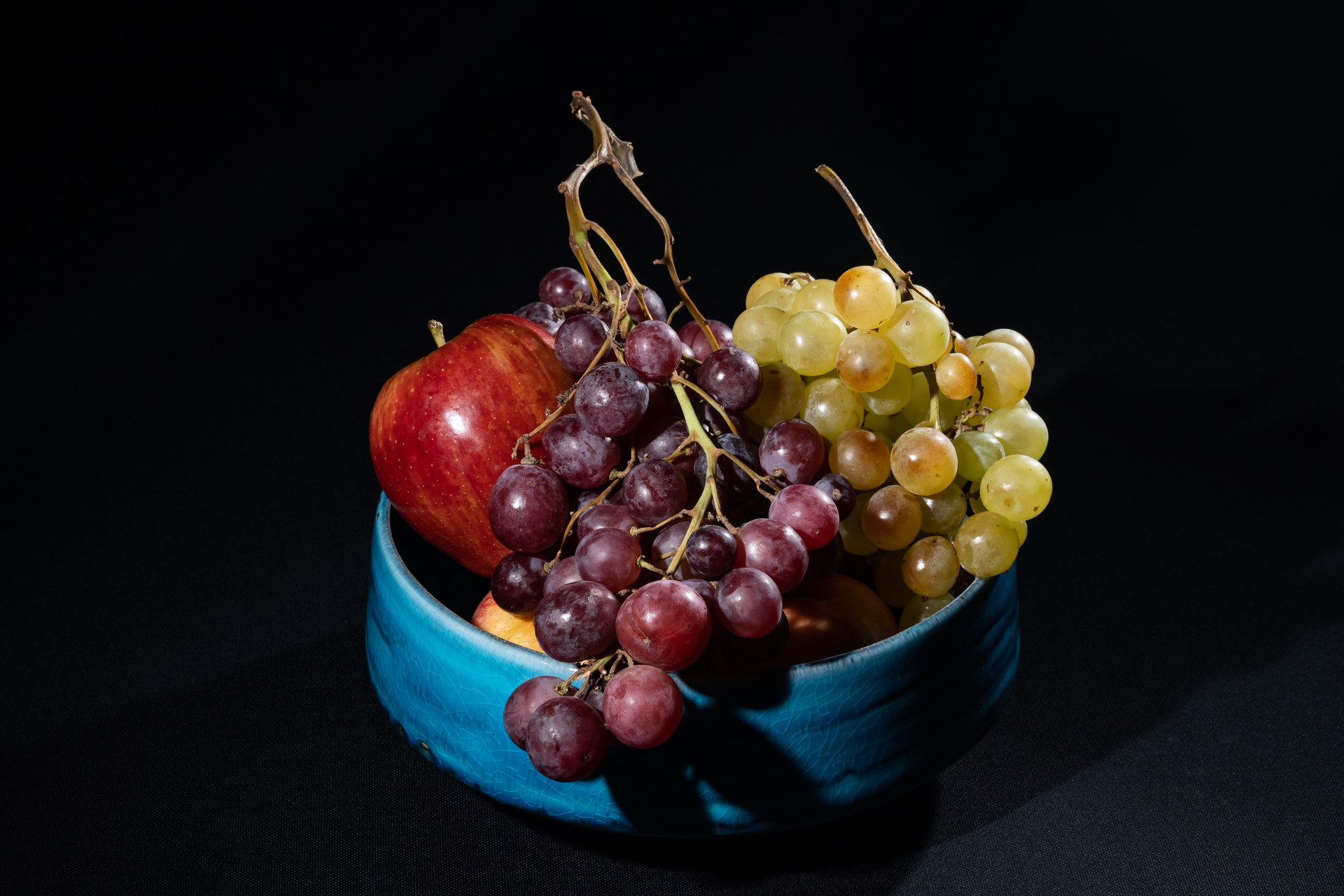 A colorful still life of fresh grapes and an apple in a blue bowl on a dark background.