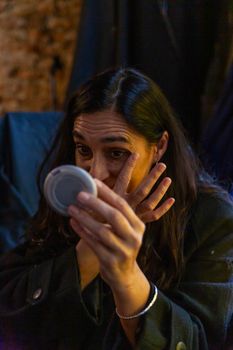 A woman applies makeup using a compact mirror in a dimly lit indoor setting.