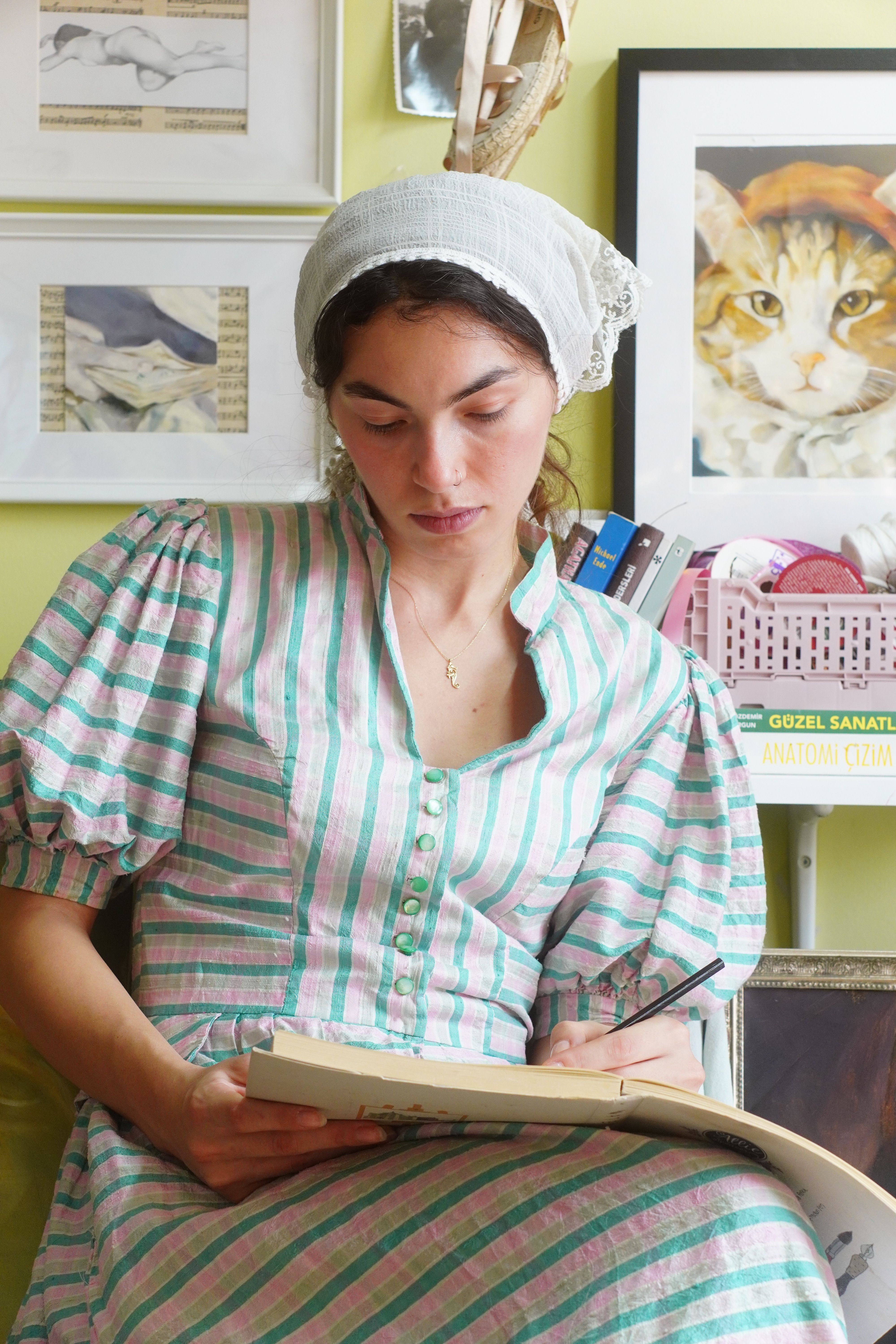 A woman in vintage attire sketches indoors beside framed art and books.