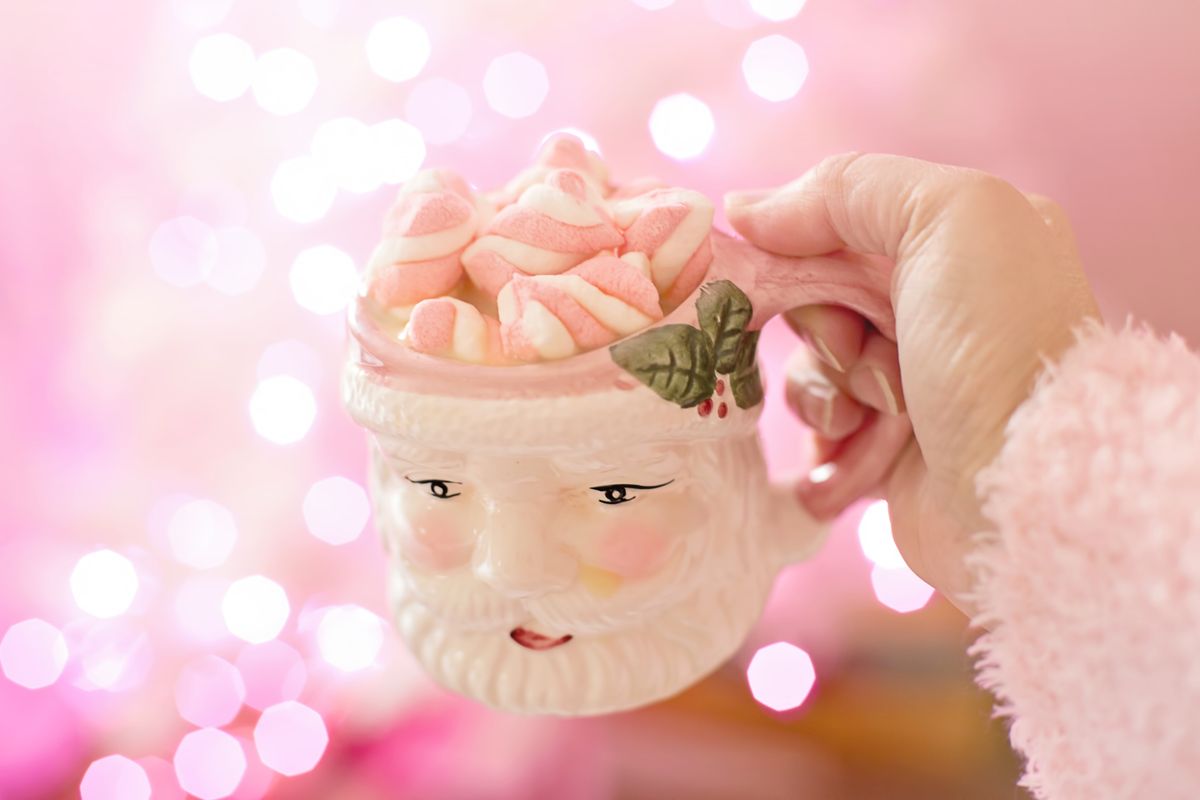 Close-up of a hand holding a Santa mug filled with pink marshmallows against a festive pink bokeh background.