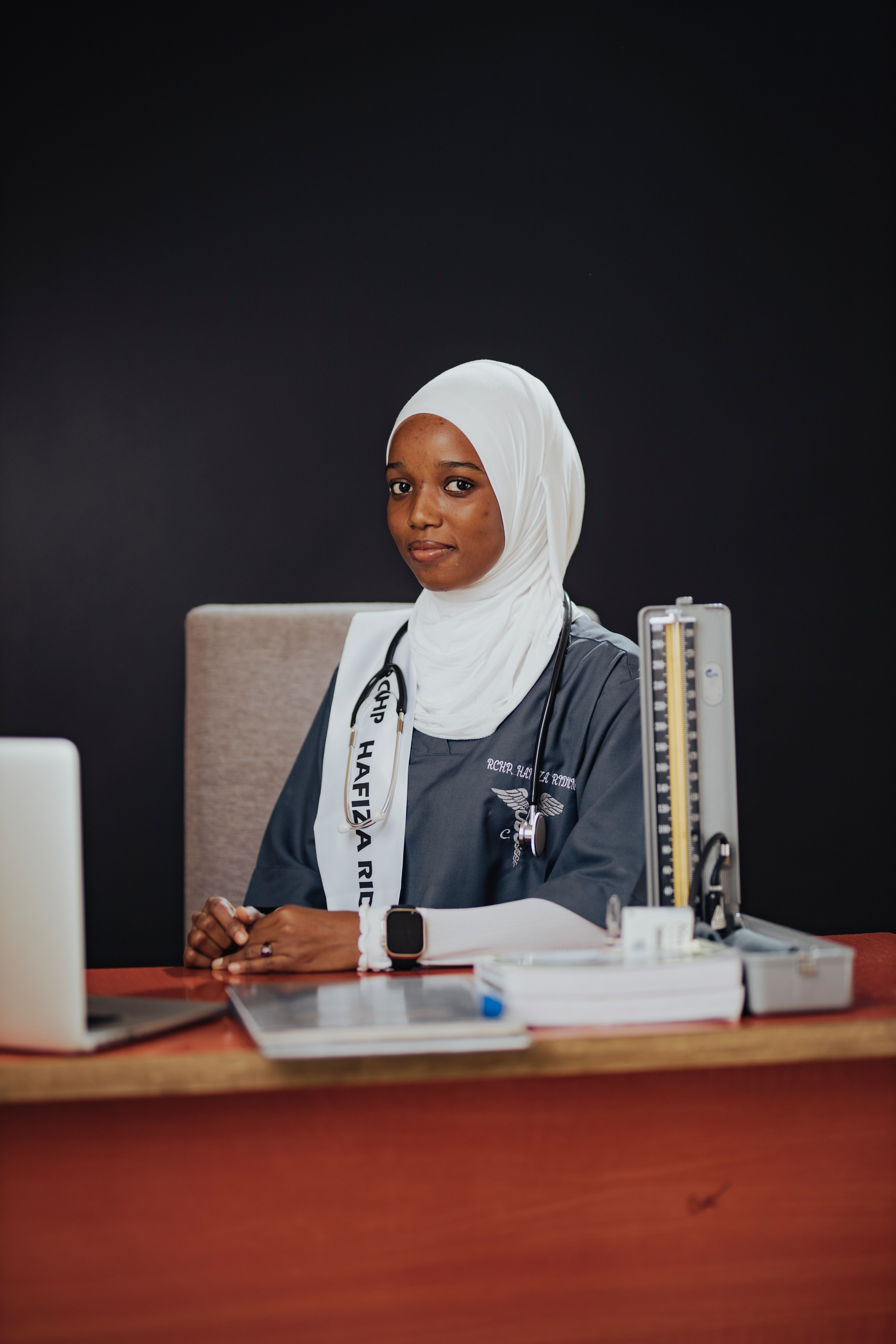 Portrait of a young female doctor wearing a stethoscope and graduation attire, confidently seated at a desk.