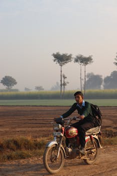 A young man riding a motorcycle on a rural road with misty fields in the background.