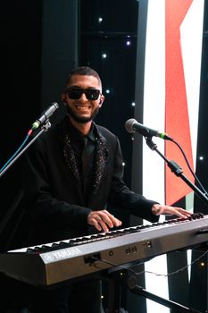 A smiling musician performing on stage with a keyboard and microphones, exuding confidence and joy.