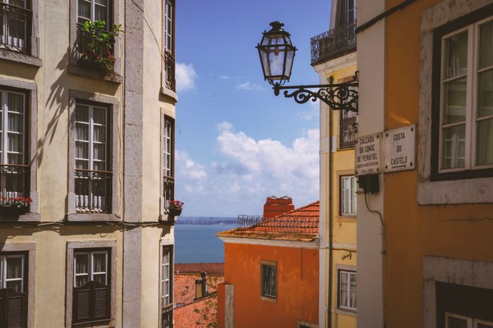 Wrought-iron street lamp and tiled façades framing a view of the Tagus from the Alfama district — Sandra Mosconi on Pexels