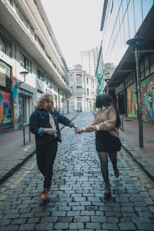 Free A young couple joyfully running and holding hands on a cobbled urban street lined with colorful graffiti. Stock Photo