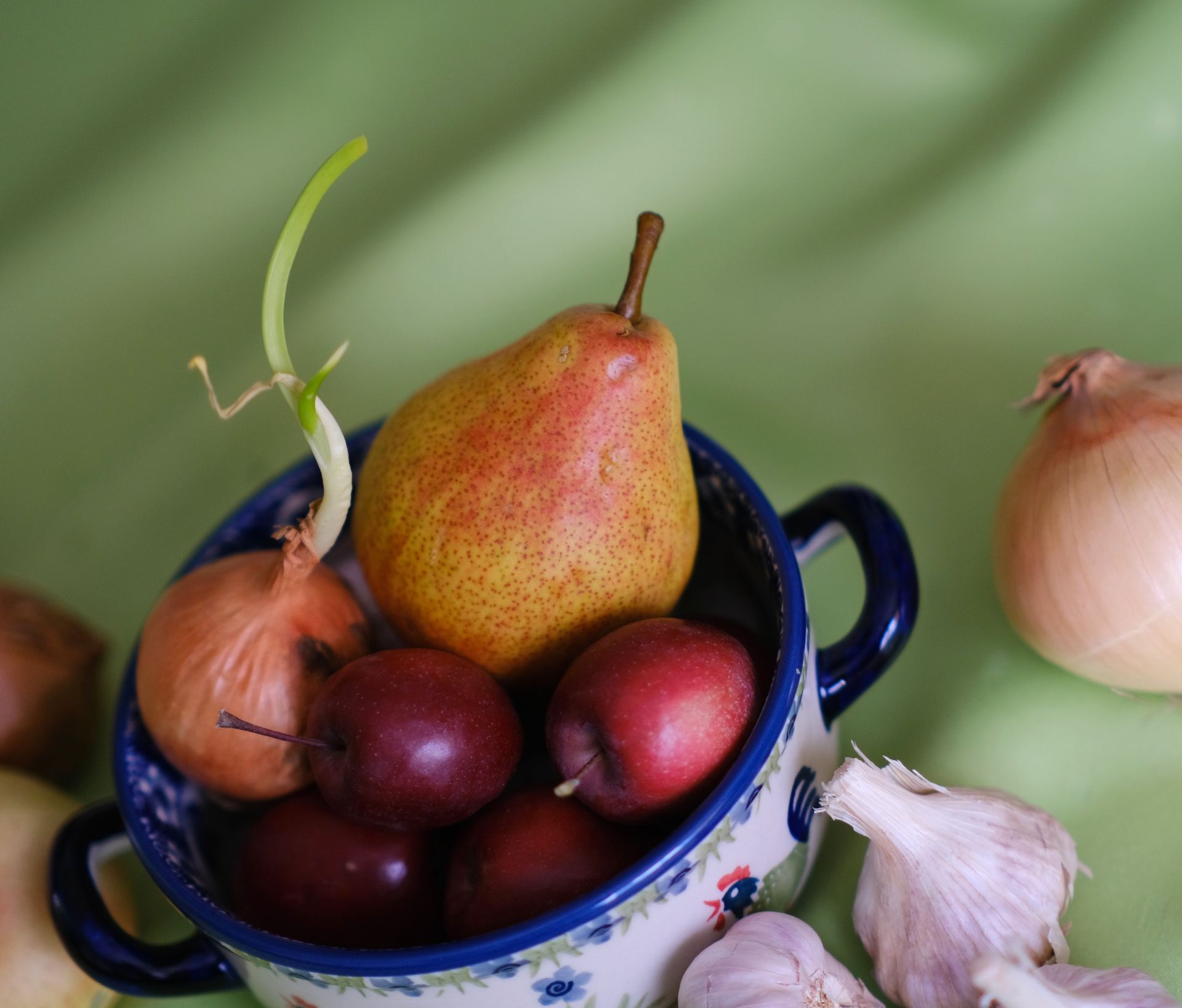 A vibrant arrangement of pear, plums, onions, and garlic in a decorative bowl.