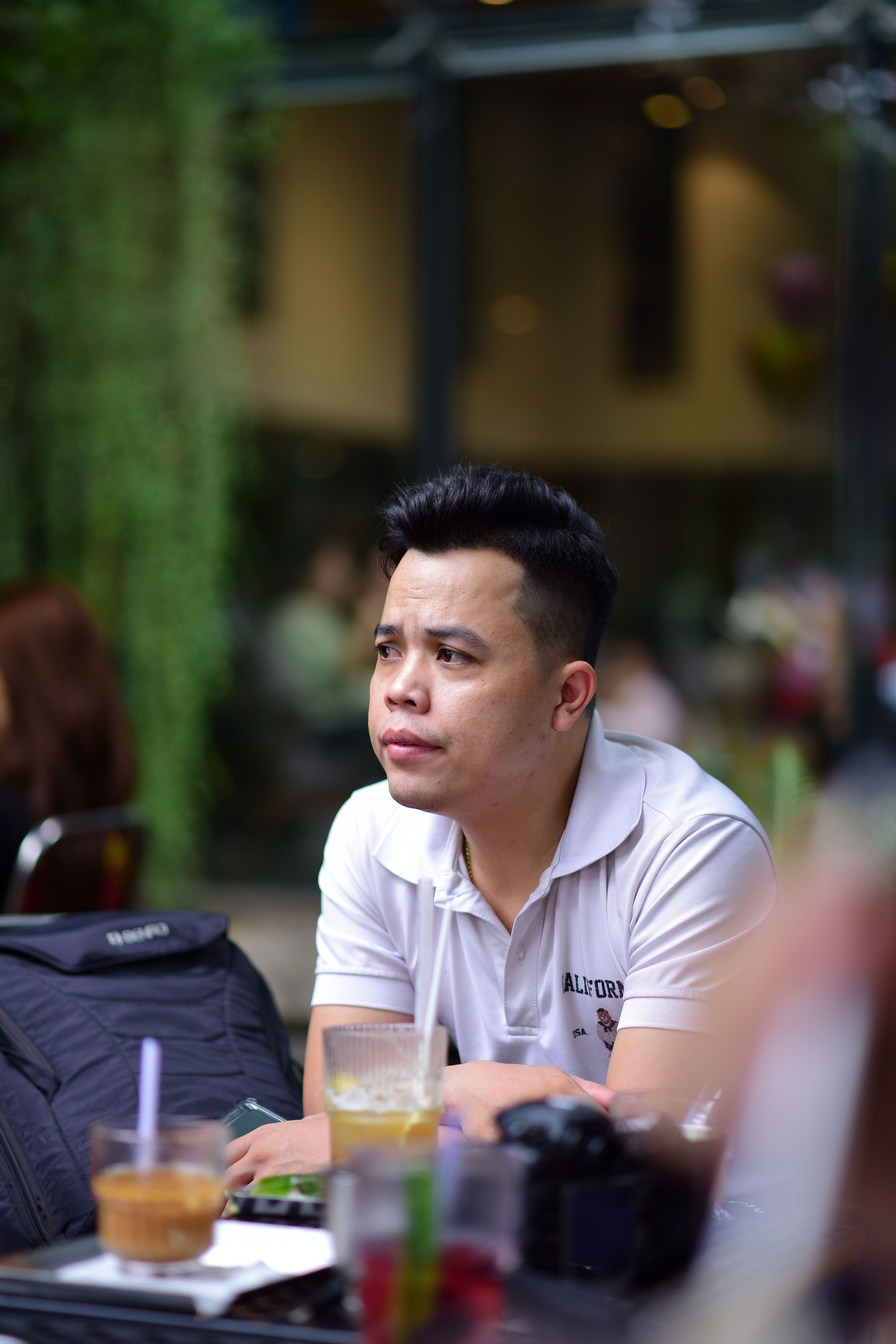 An adult man in a white shirt sits pensively at an outdoor café surrounded by drinks and a camera.