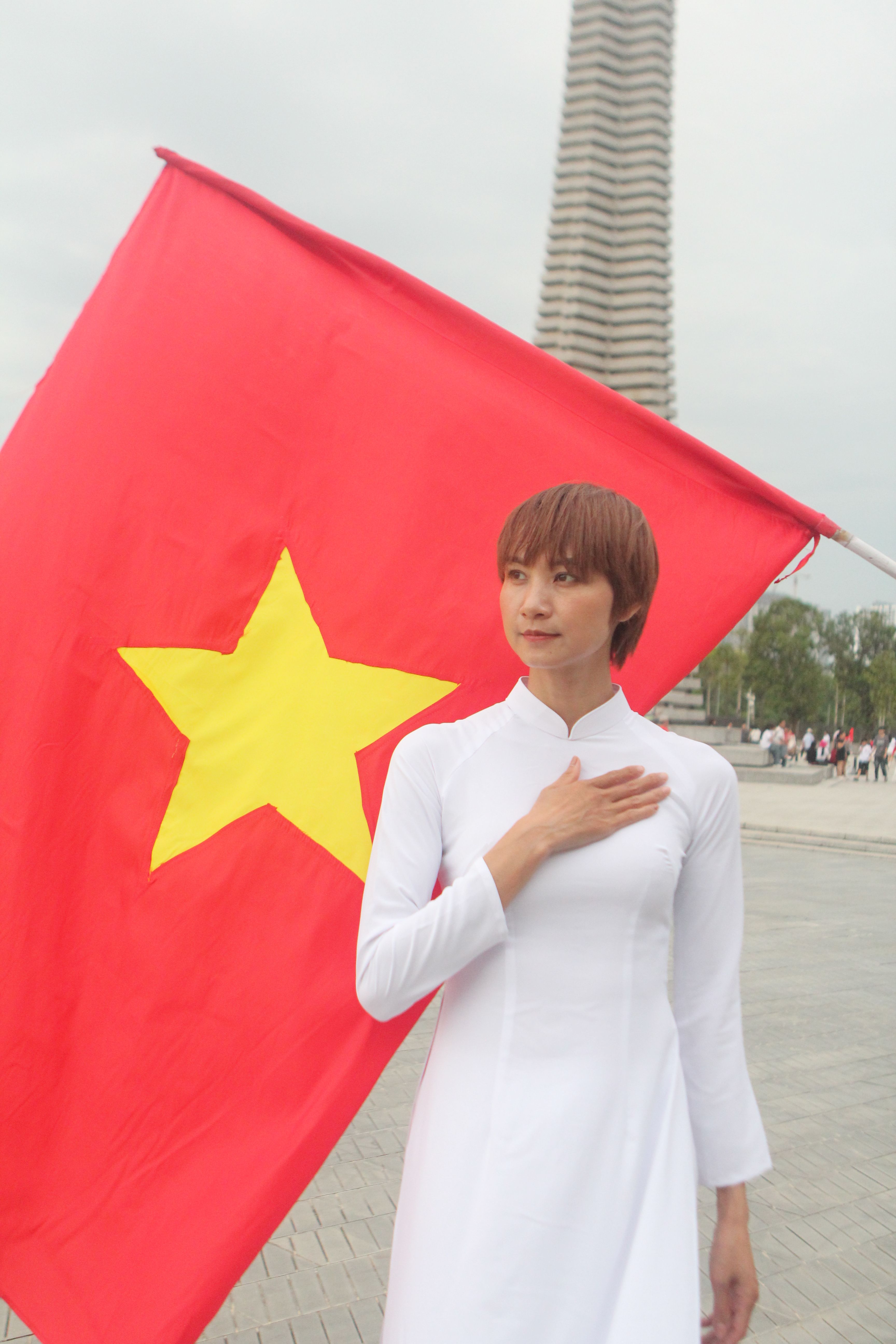 A woman in traditional attire stands with a Vietnamese flag, showing patriotism in an outdoor setting.