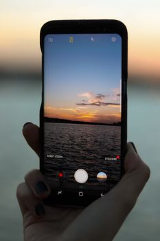 Hand holding smartphone, photographing sunset over a calm lake.