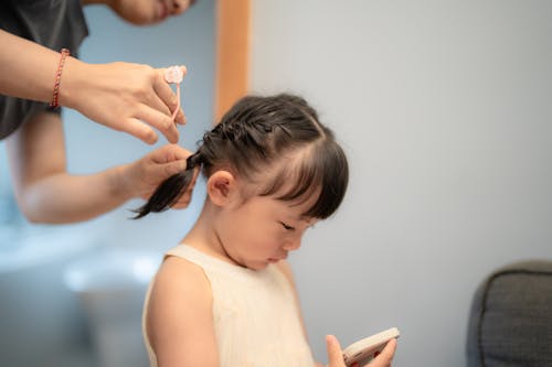 A young girl with braided hair looks at a smartphone indoors while receiving hair styling assistance.