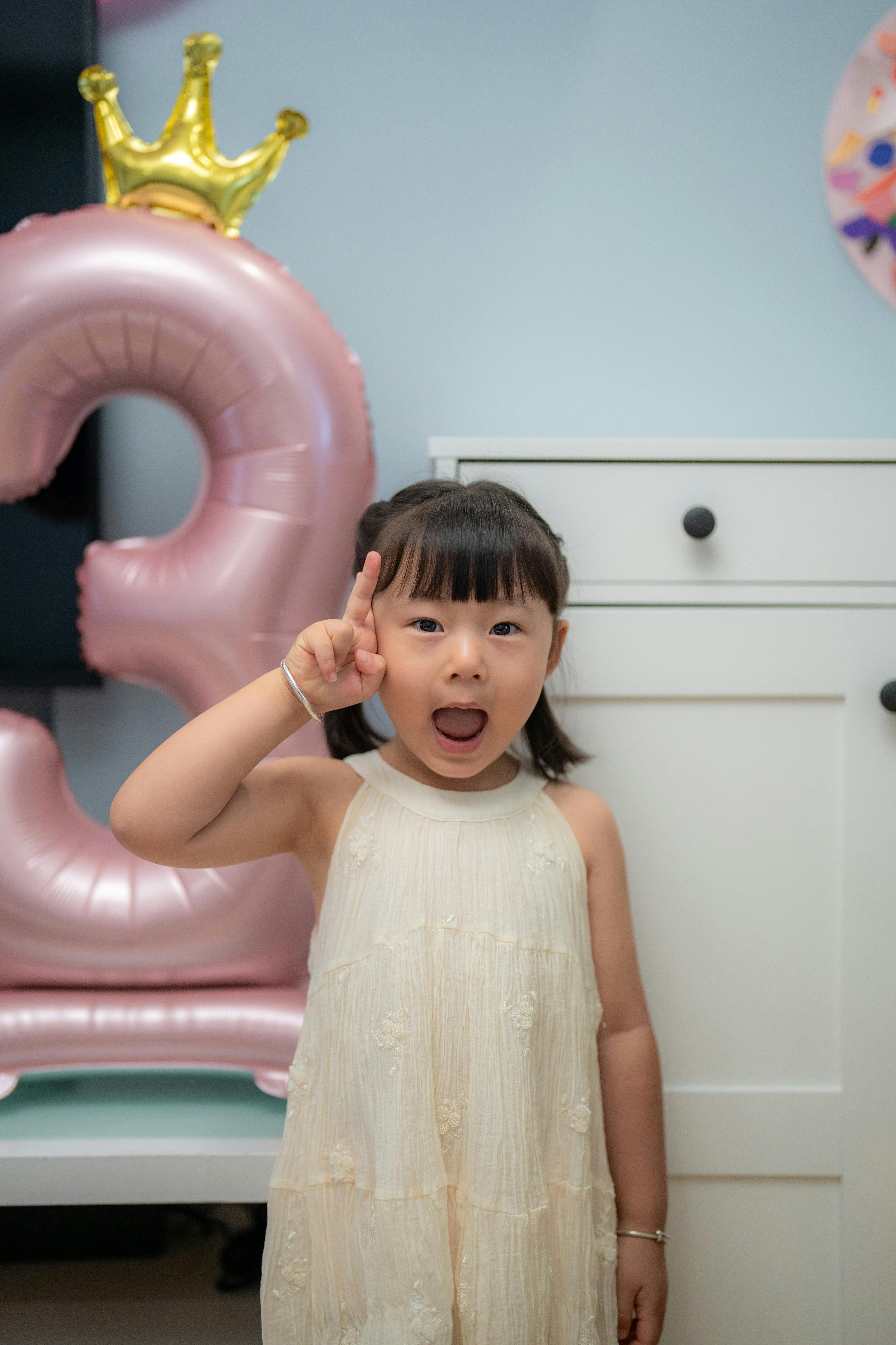 Cute Asian girl celebrating her third birthday with a party indoors, posing happily with a pink balloon.