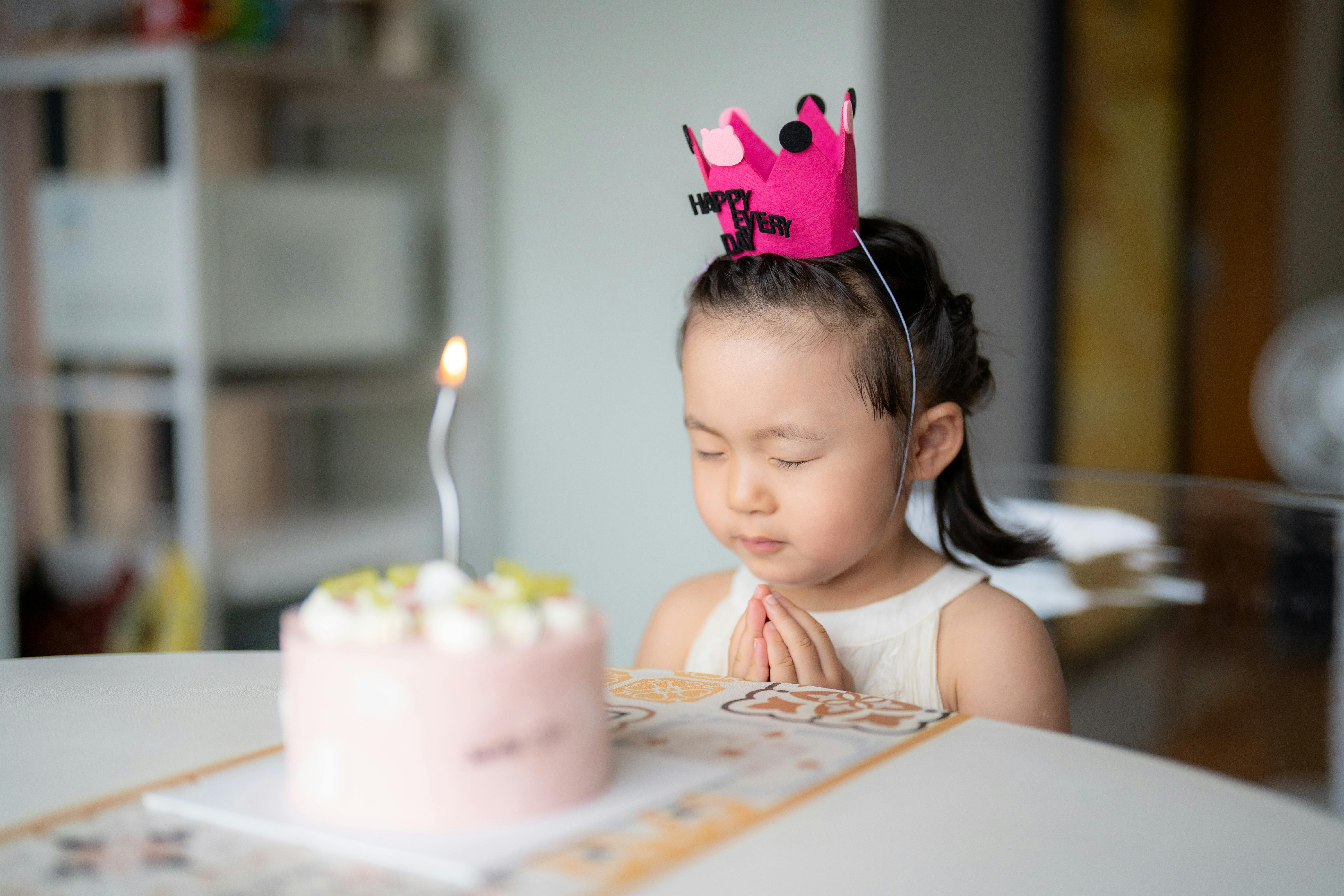 A young girl celebrates her birthday with a delightful pink cake and lit candle, wearing a festive crown.