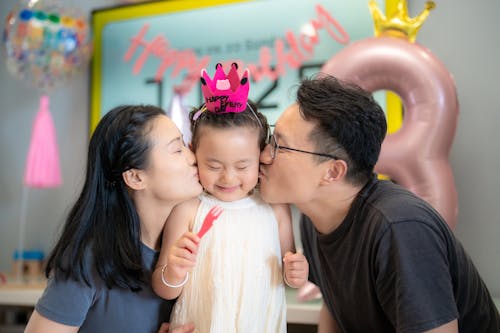 A cheerful moment of parents kissing their child during a birthday party indoors.