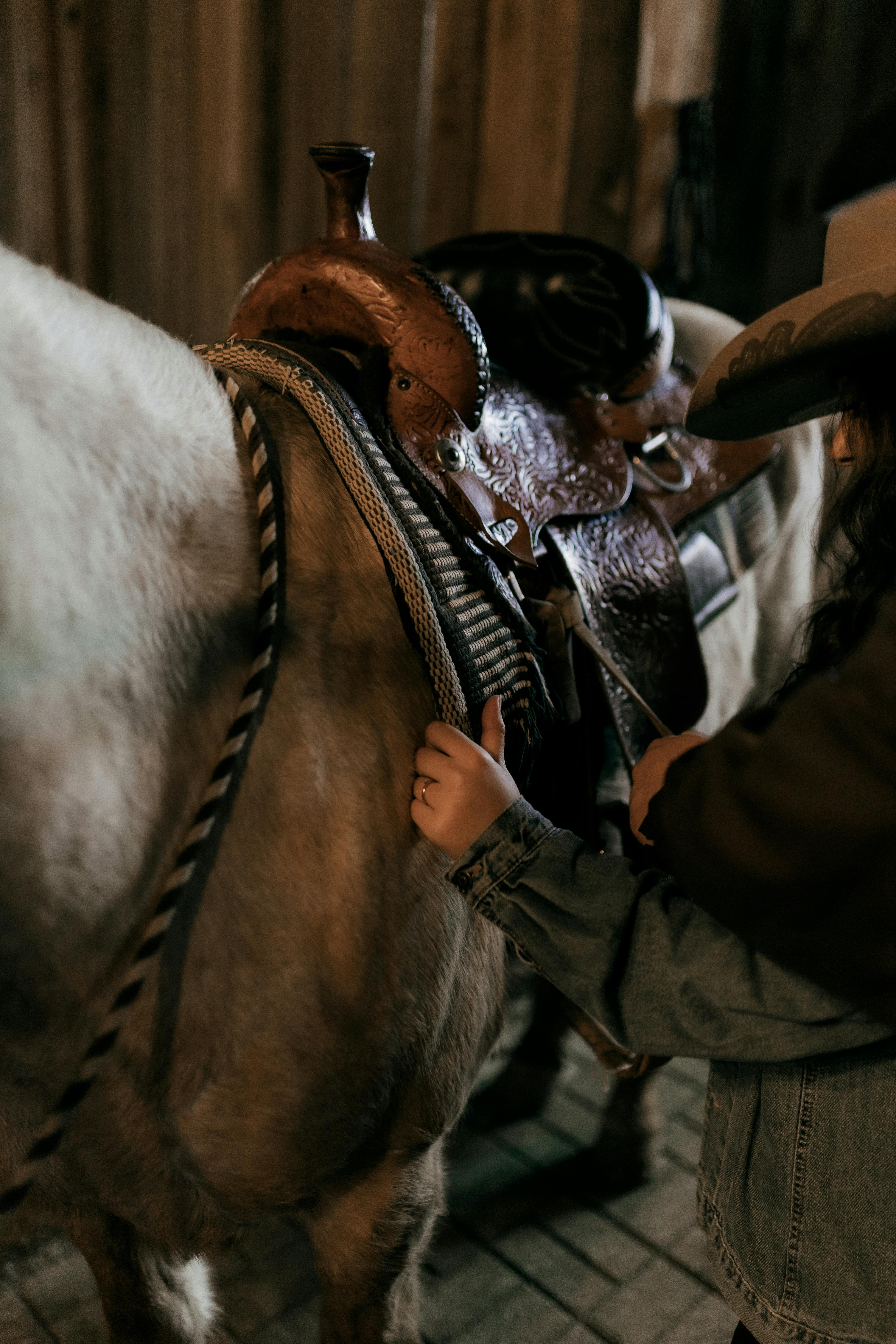 An individual in a cowboy hat adjusting a saddle on a horse inside a dimly lit barn.
