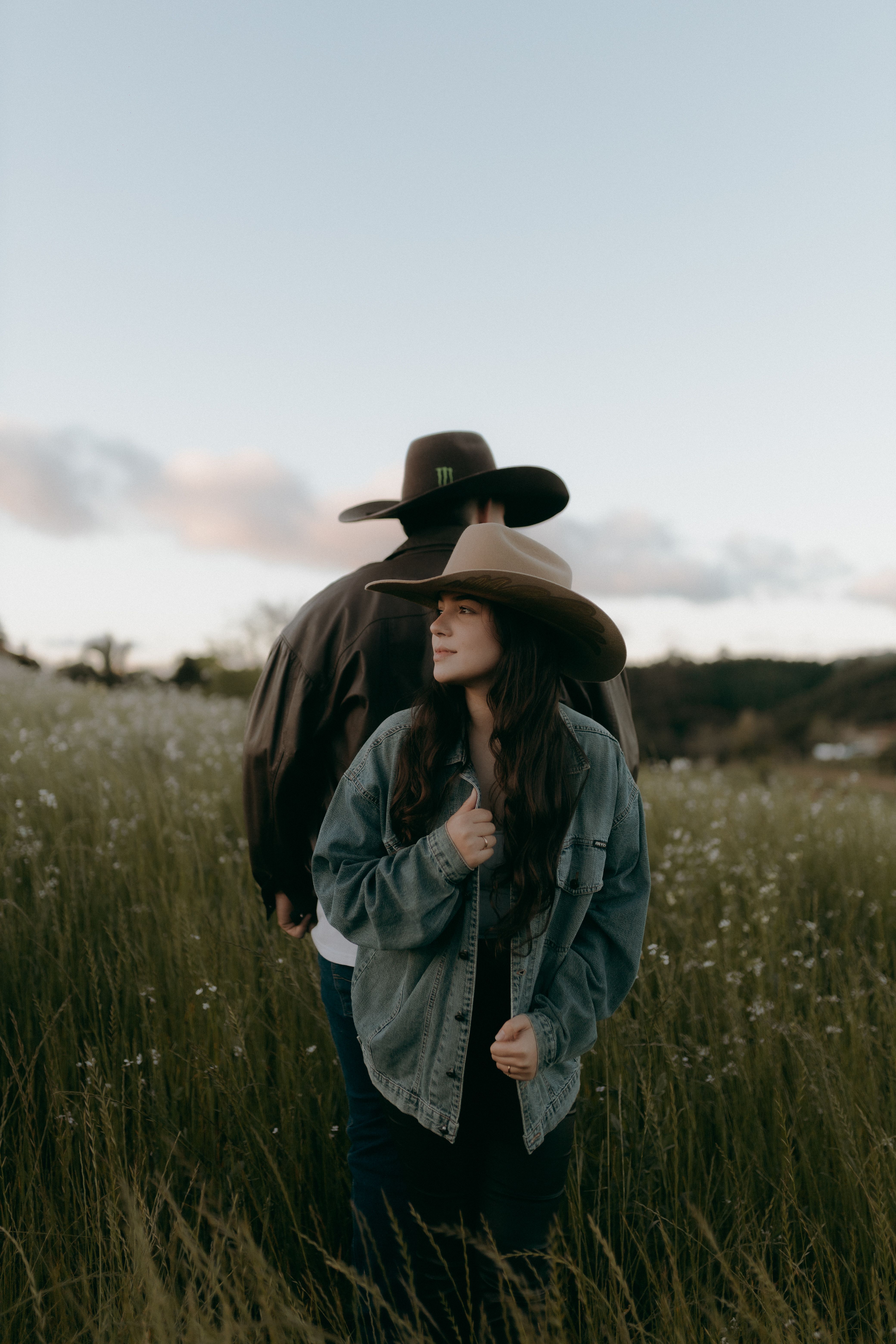Two people wearing cowboy hats stand in a scenic field during a serene day.