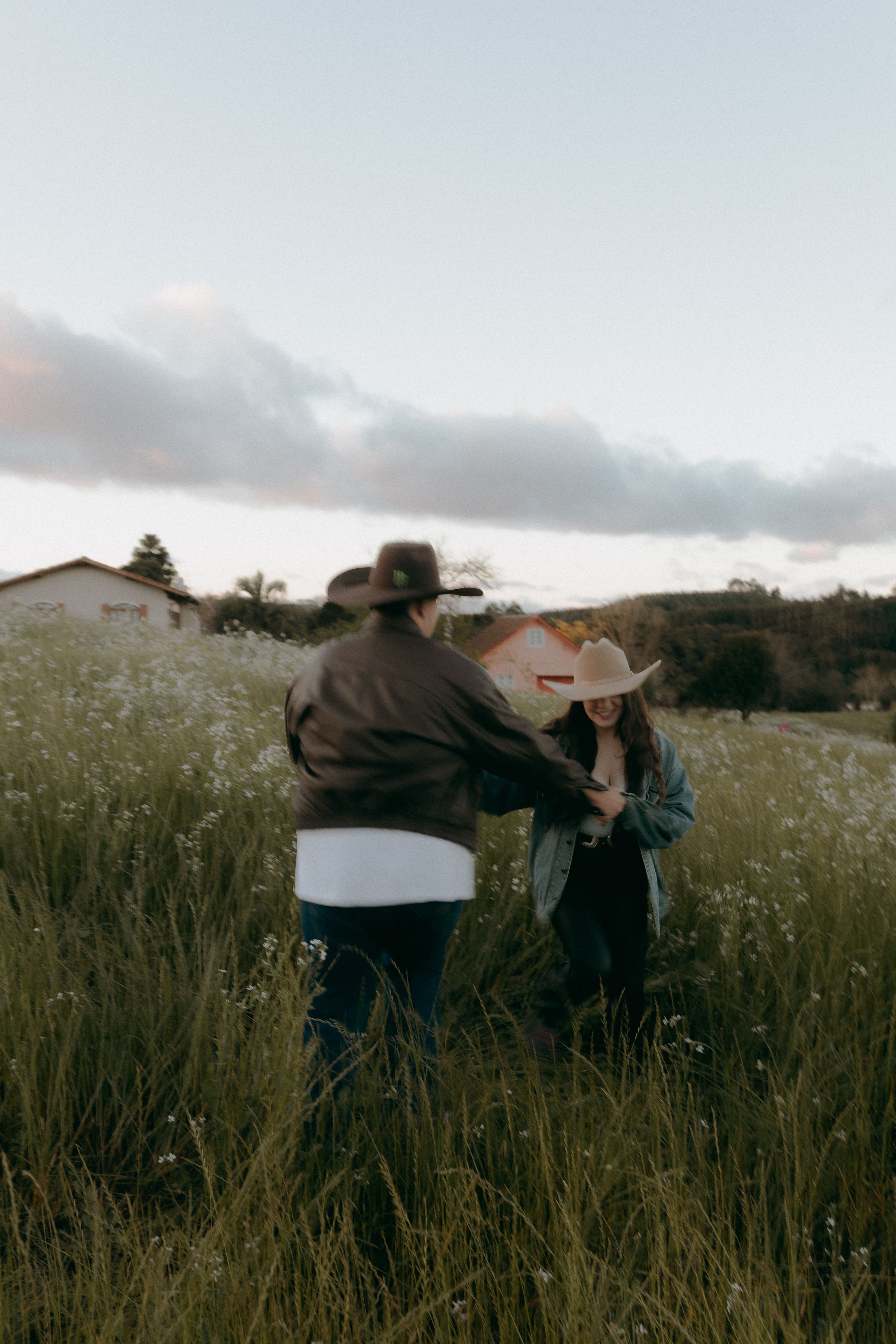A joyful couple in cowboy hats walking through a picturesque field during sunset.